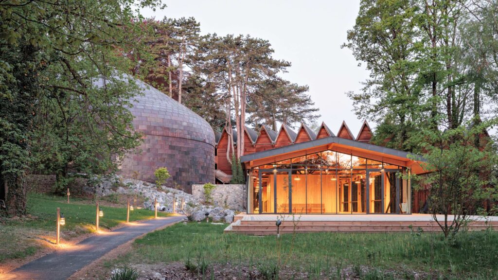 A modern building with a large copper-colored dome and a glass-walled pavilion, nestled among trees.