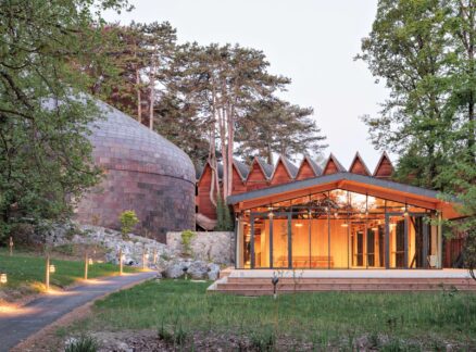 A modern building with a large copper-colored dome and a glass-walled pavilion, nestled among trees.