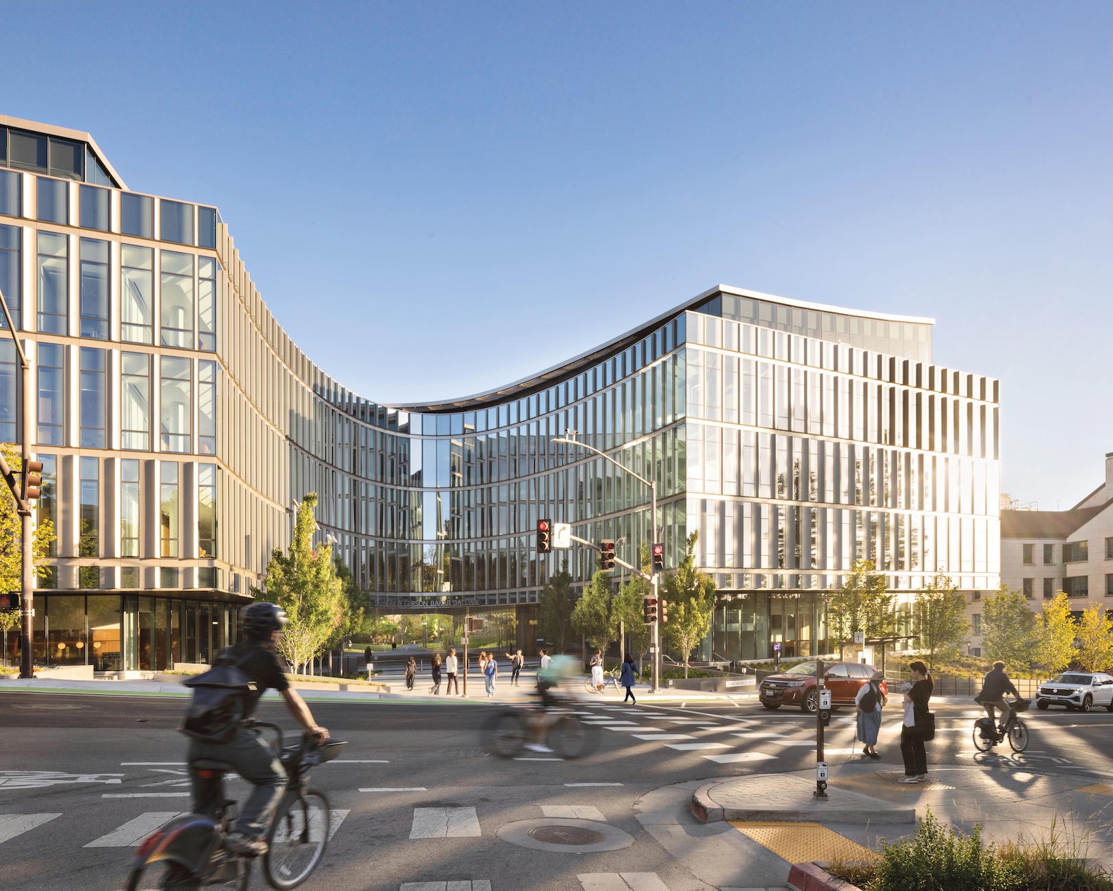 A curved, multi-story glass office building near the Lucas Museum of Narrative Art on a sunny day, with pedestrians and cyclists crossing a city intersection in the foreground.