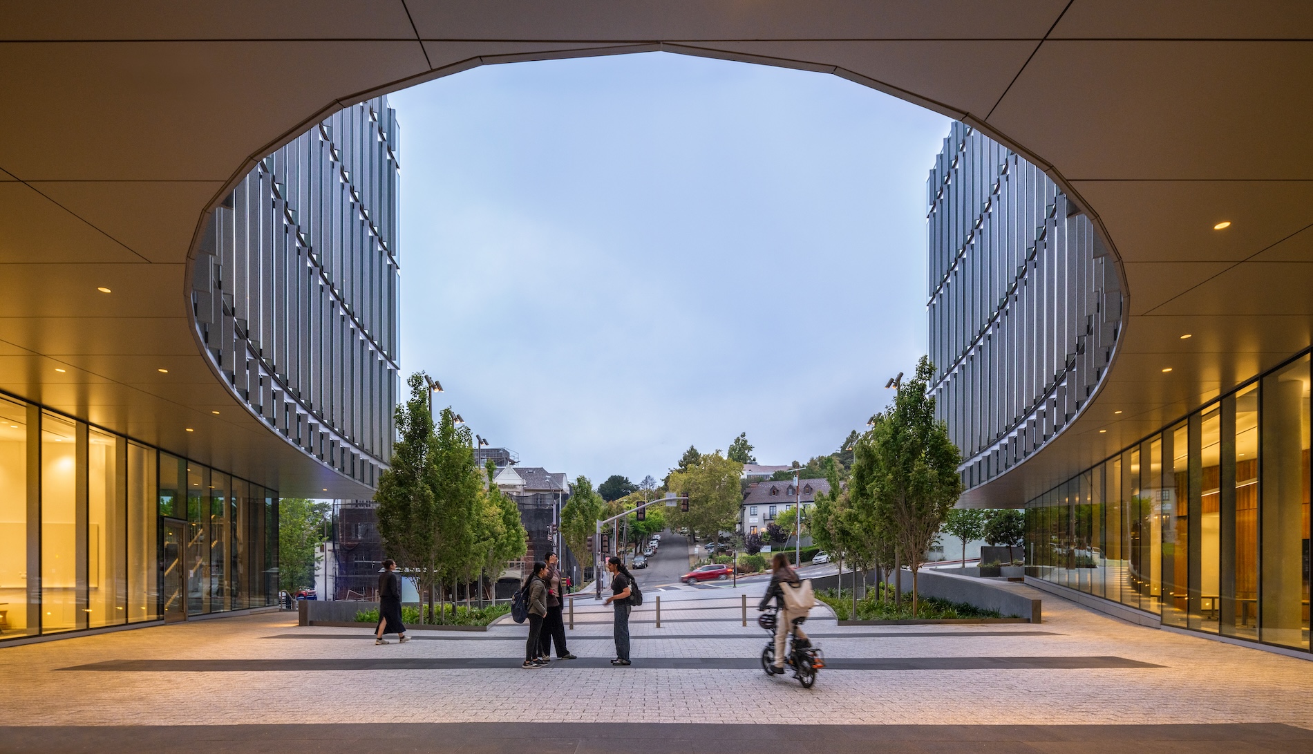 People walk and cycle through a paved plaza between the modern glass buildings of the Lucas Museum of Narrative Art, beneath a large, circular architectural cutout.