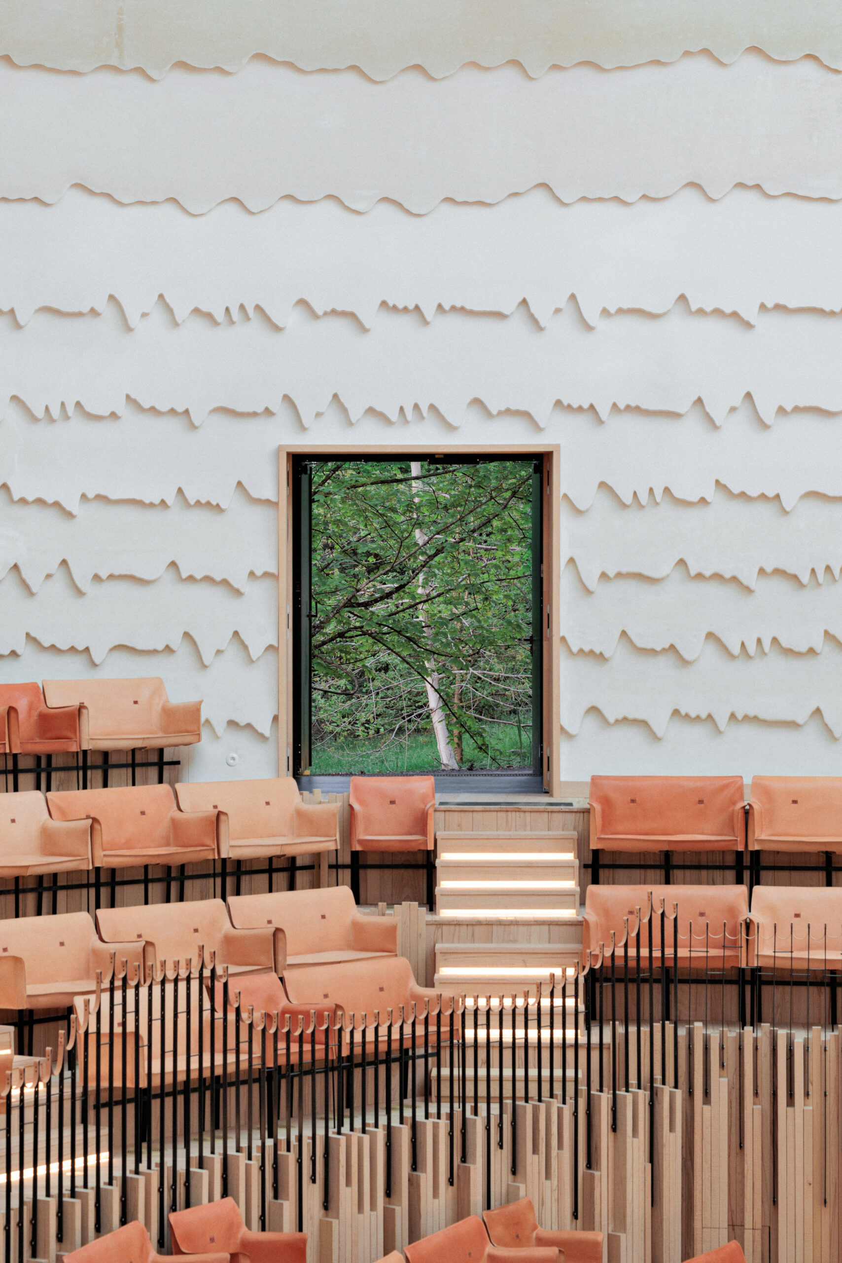 Inside La Source by PCA-STREAM, a modern concert hall with white textural walls with a melting quality, earth-tone upholstery and a window into the green outdoors.
