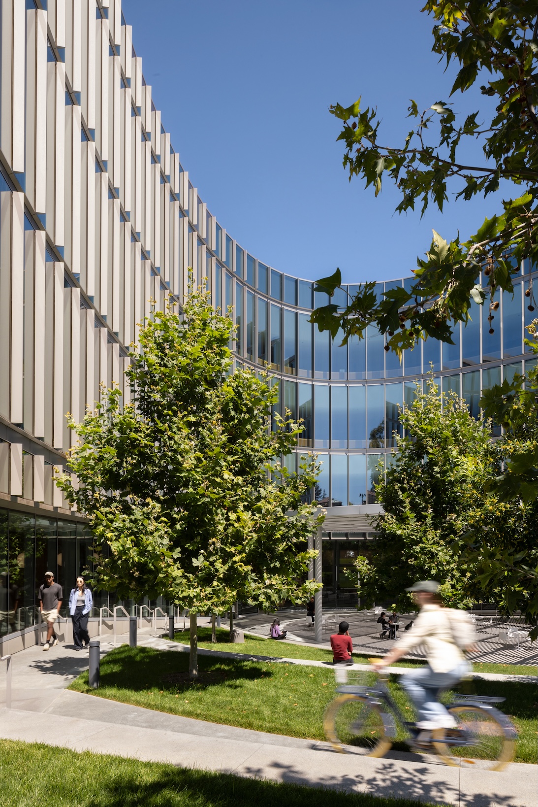 The curved, multi-story glass facade of the Lucas Museum of Narrative Art features vertical metal louvers overlooking a landscaped courtyard with trees and people.