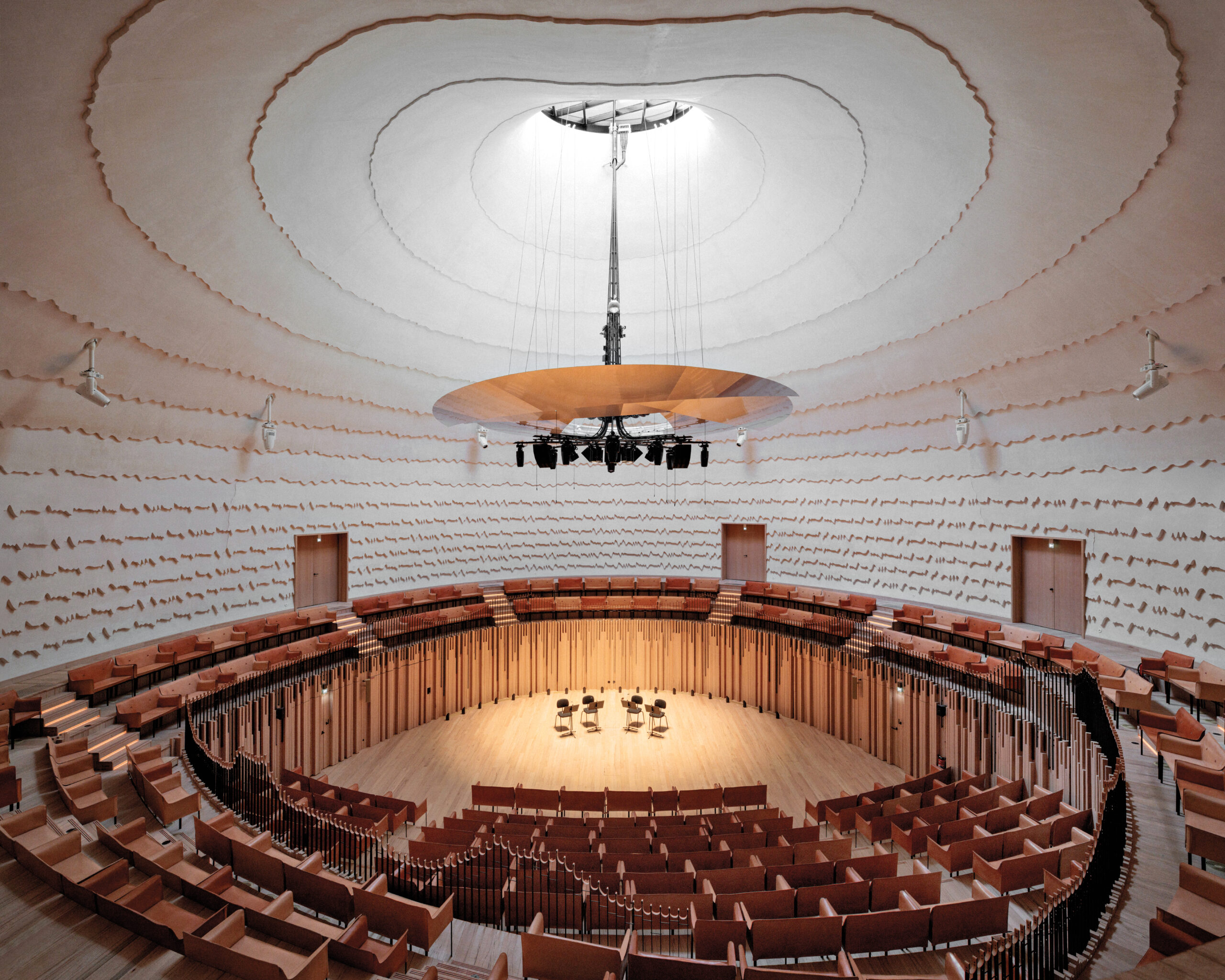 Inside La Source by PCA-STREAM, a modern concert hall with white textural walls and a sweeping view of a central stage underneath a skylight.