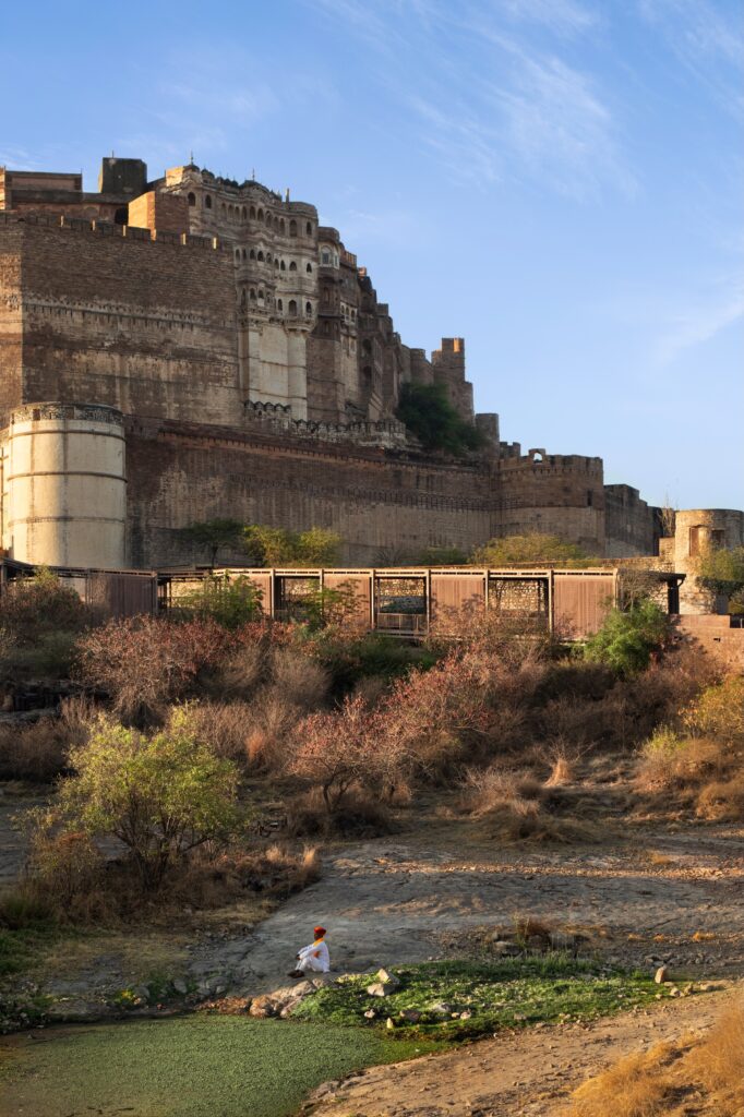 Mehrangarh Fort towers above a rocky landscape, reminiscent of the Barbara and Gerson Bakar Gateway, while a person in traditional clothing sits by a small pond.