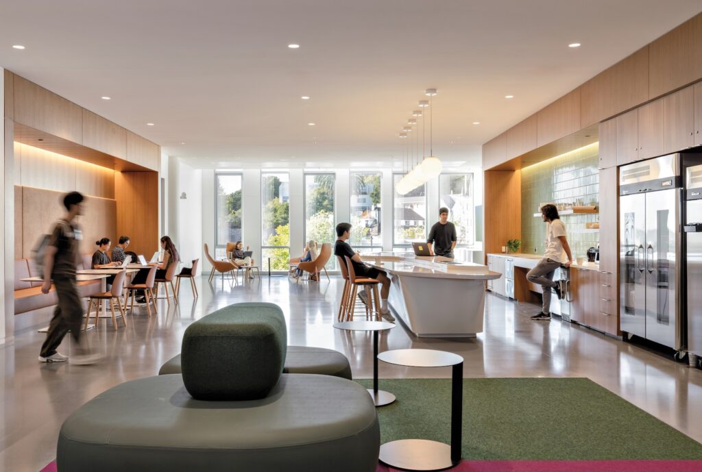 People work and socialize in a modern, open-plan office lounge that evokes the creative atmosphere of the Lucas Museum of Narrative Art, featuring tables, a kitchen island, and a green seating area.
