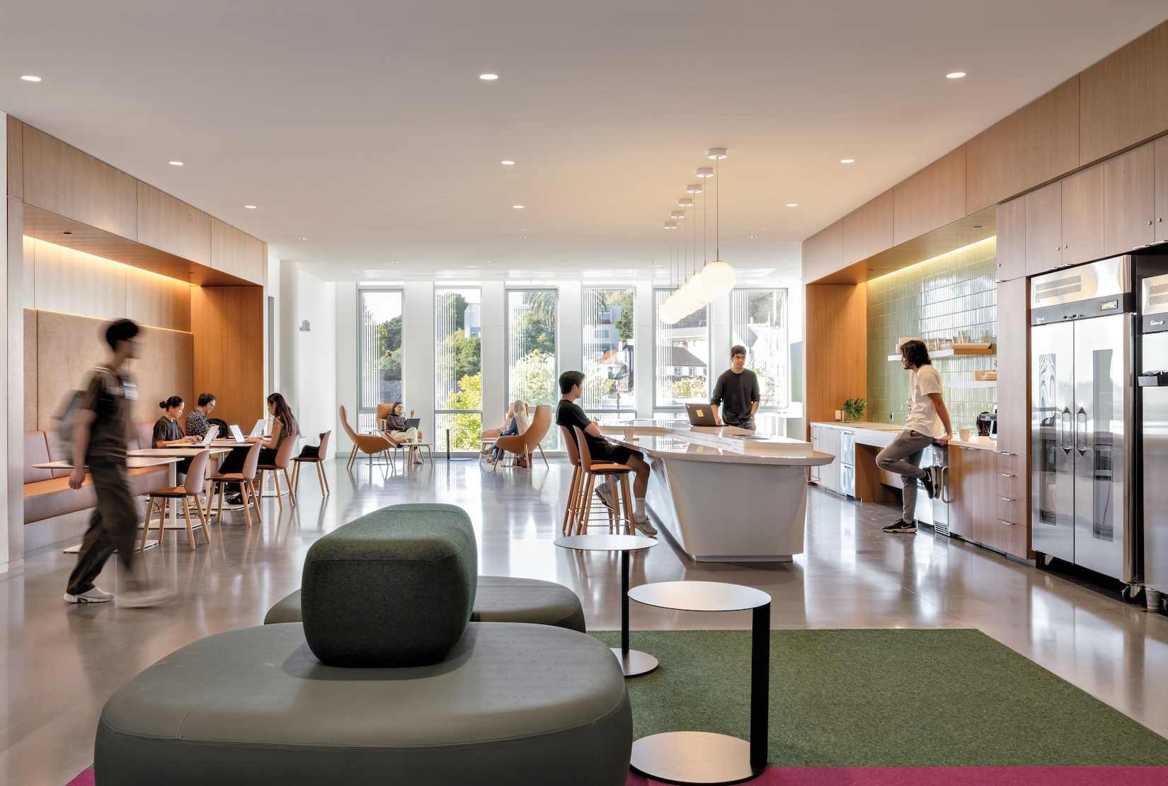 People work and socialize in a modern, open-plan office lounge that evokes the creative atmosphere of the Lucas Museum of Narrative Art, featuring tables, a kitchen island, and a green seating area.