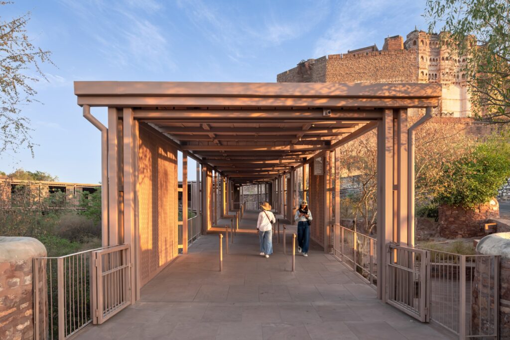 Two people walk through the modern, metal Barbara and Gerson Bakar Gateway at UC Berkeley, leading toward a stone fortress.