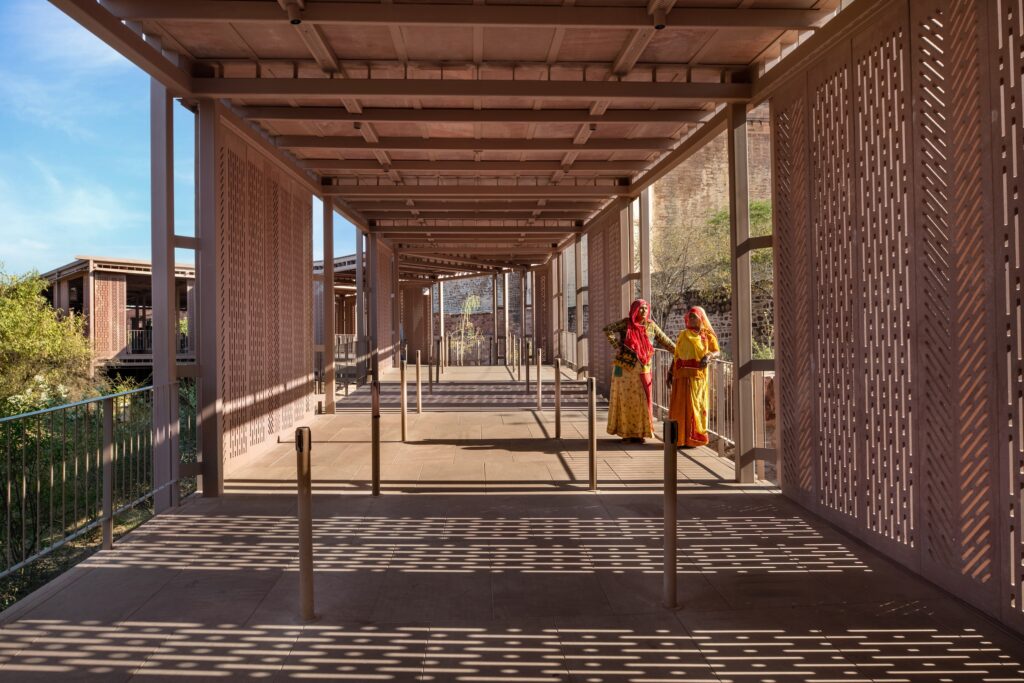 Two women in traditional vibrant saris stand on the covered stone walkway of the Barbara and Gerson Bakar Gateway at UC Berkeley, framed by patterned screens.