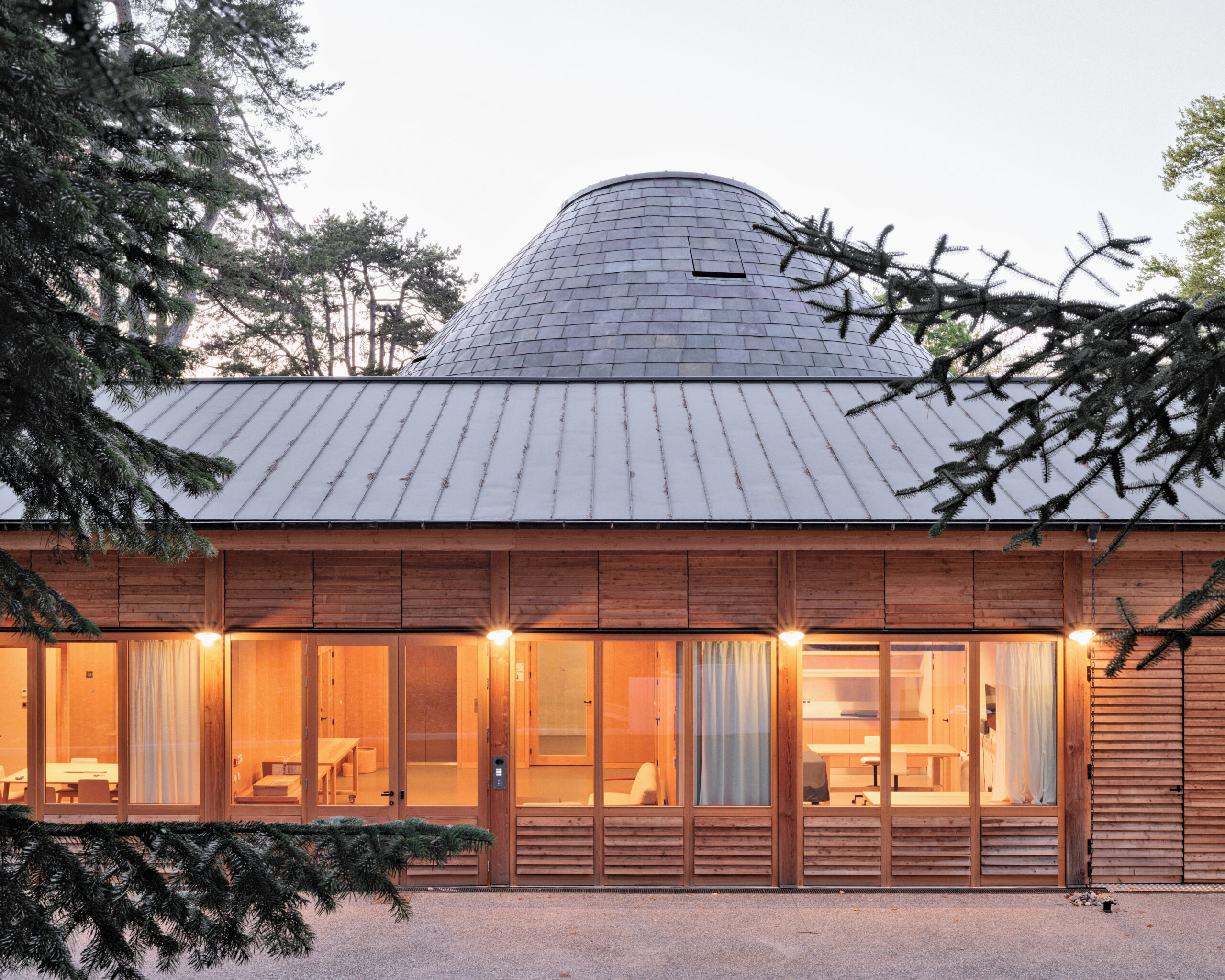 PCA-STREAM: Exterior view of La Source, with wood walls and golden lighting, and pine trees around the edges.