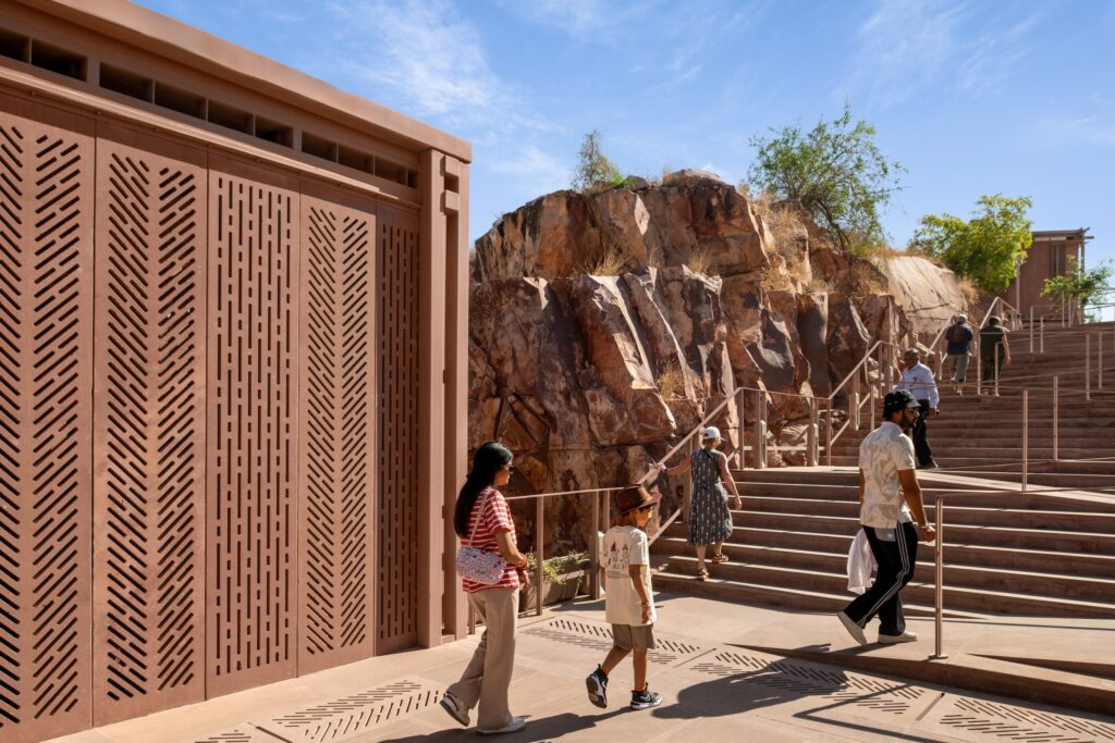 People walk up the tiered red stone staircase of the Barbara and Gerson Bakar Gateway at UC Berkeley, set alongside a large, rocky cliff face.