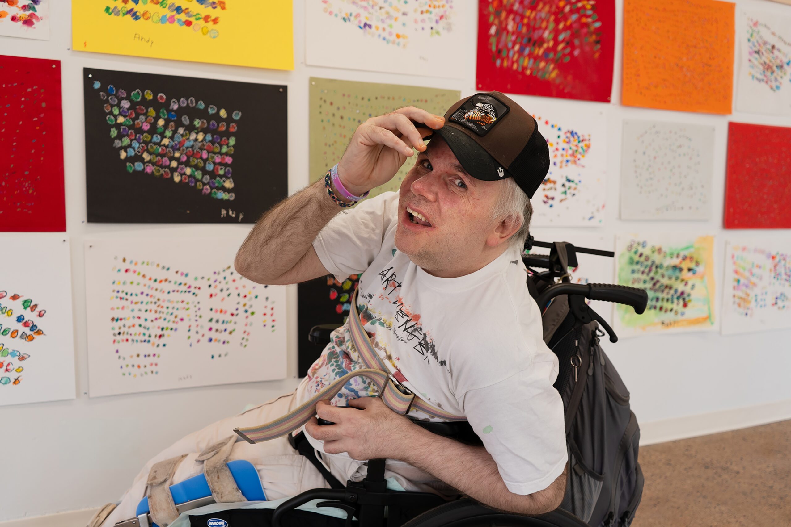A man in a wheelchair wearing a graphic t-shirt and baseball cap, posed in front of a gallery wall covered in colorful abstract art.