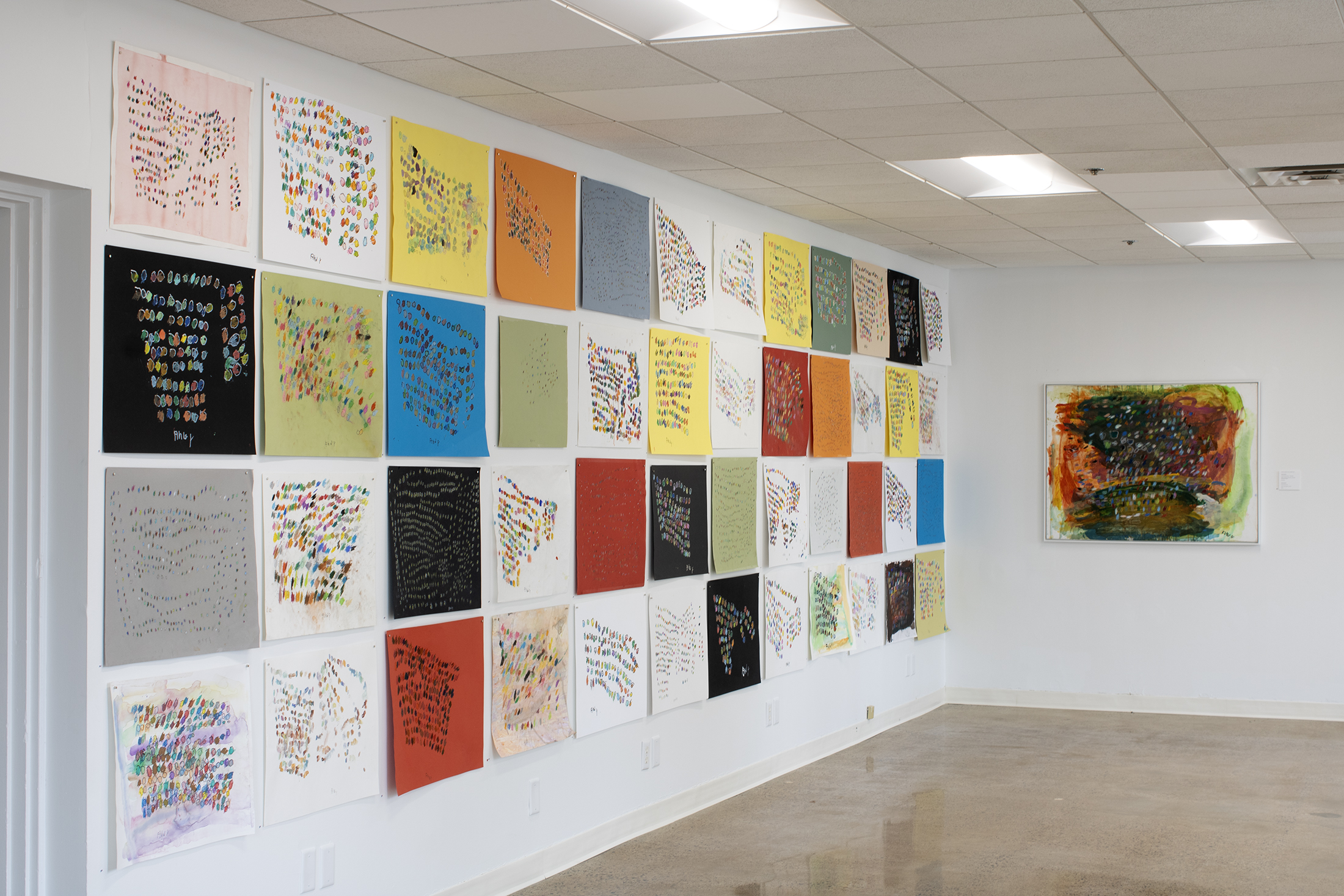 A wall in a gallery covered with a grid of small, colorful abstract drawings, with one large framed abstract painting on the adjacent wall.