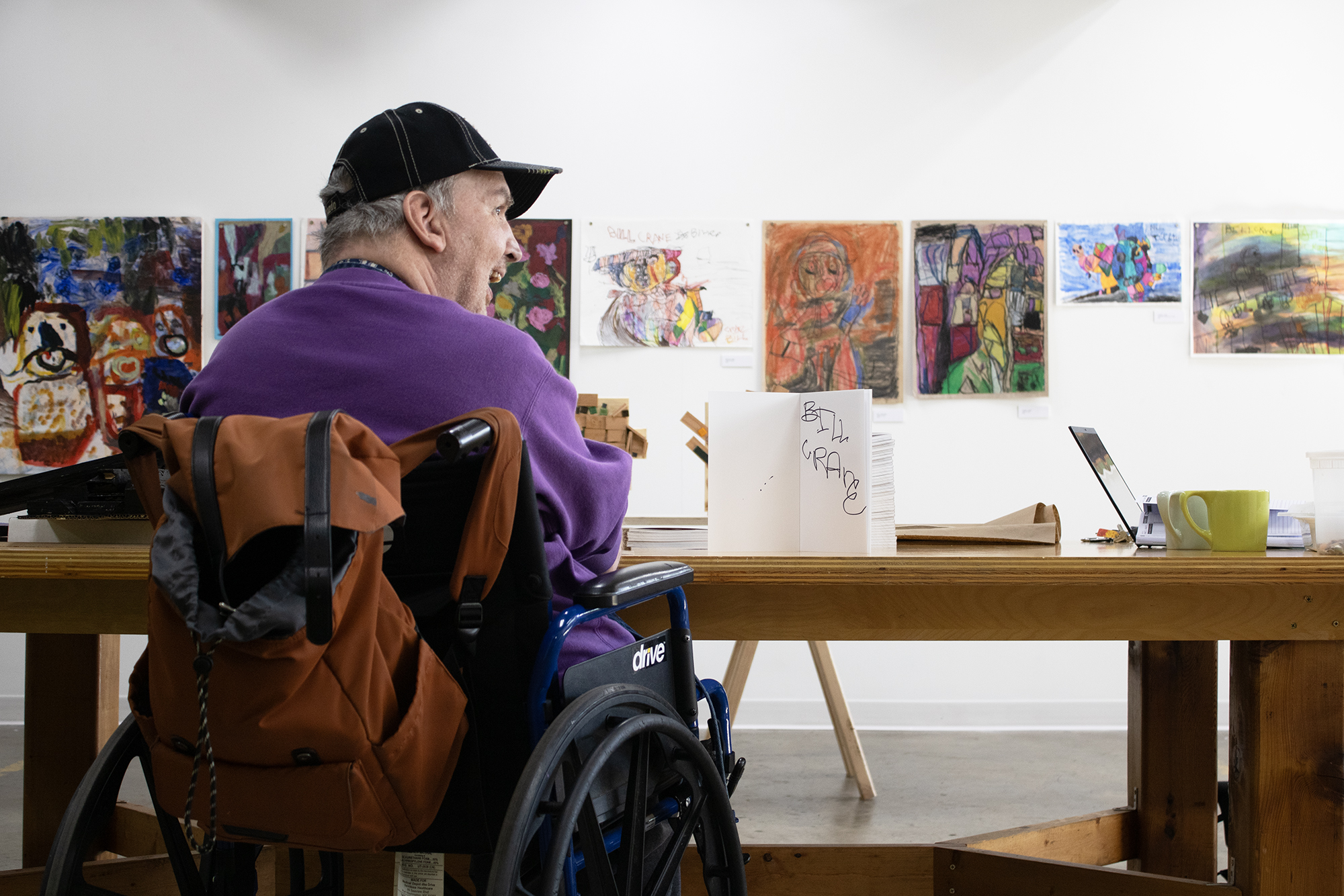 A man in a wheelchair and a purple sweater sits at a table in an art gallery, facing a book titled “BILL GRACE” displayed among various paintings on the walls.
