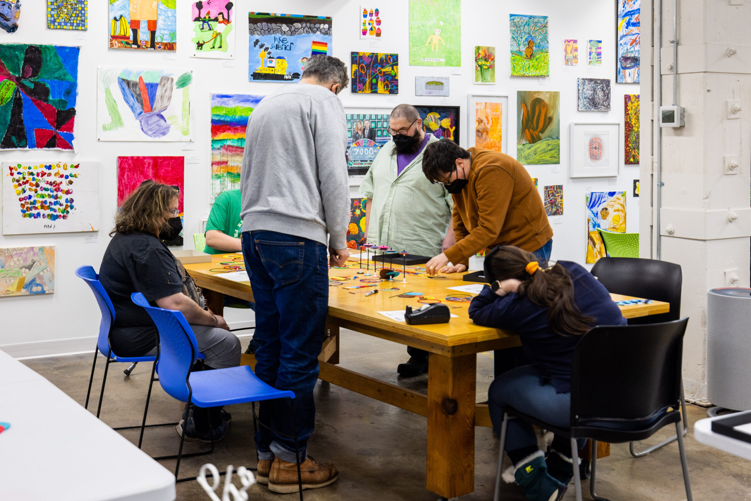 People working together at a large wooden table in an art gallery filled with paintings on the walls.