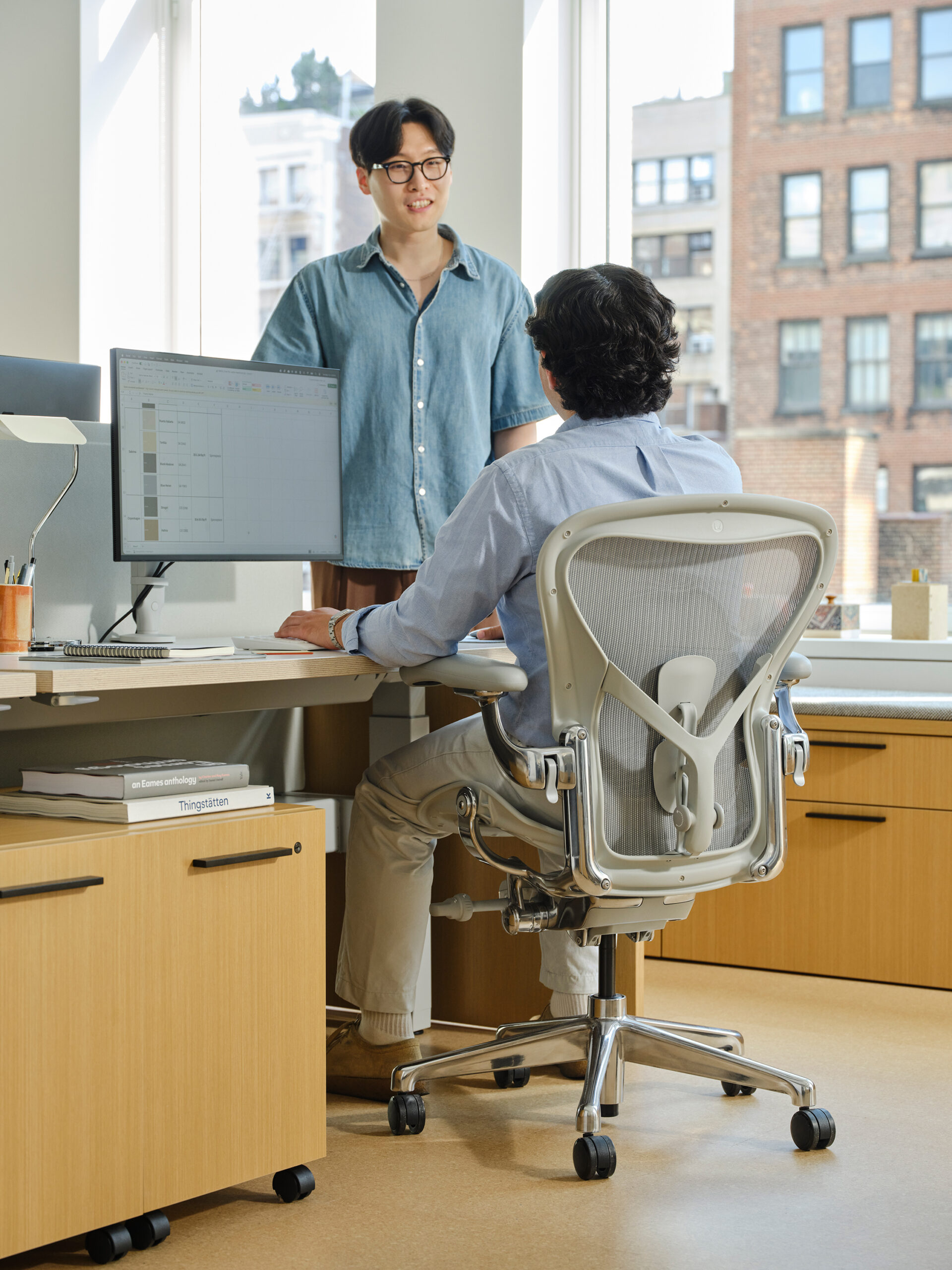 Two men in an office, one sitting at a desk with a computer and the other standing nearby.
