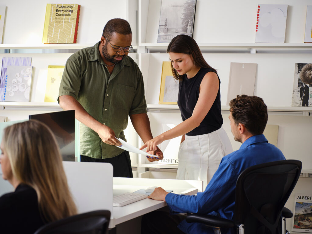 Two people review a MillerKnoll document together in an office while a third person works at a desk nearby.