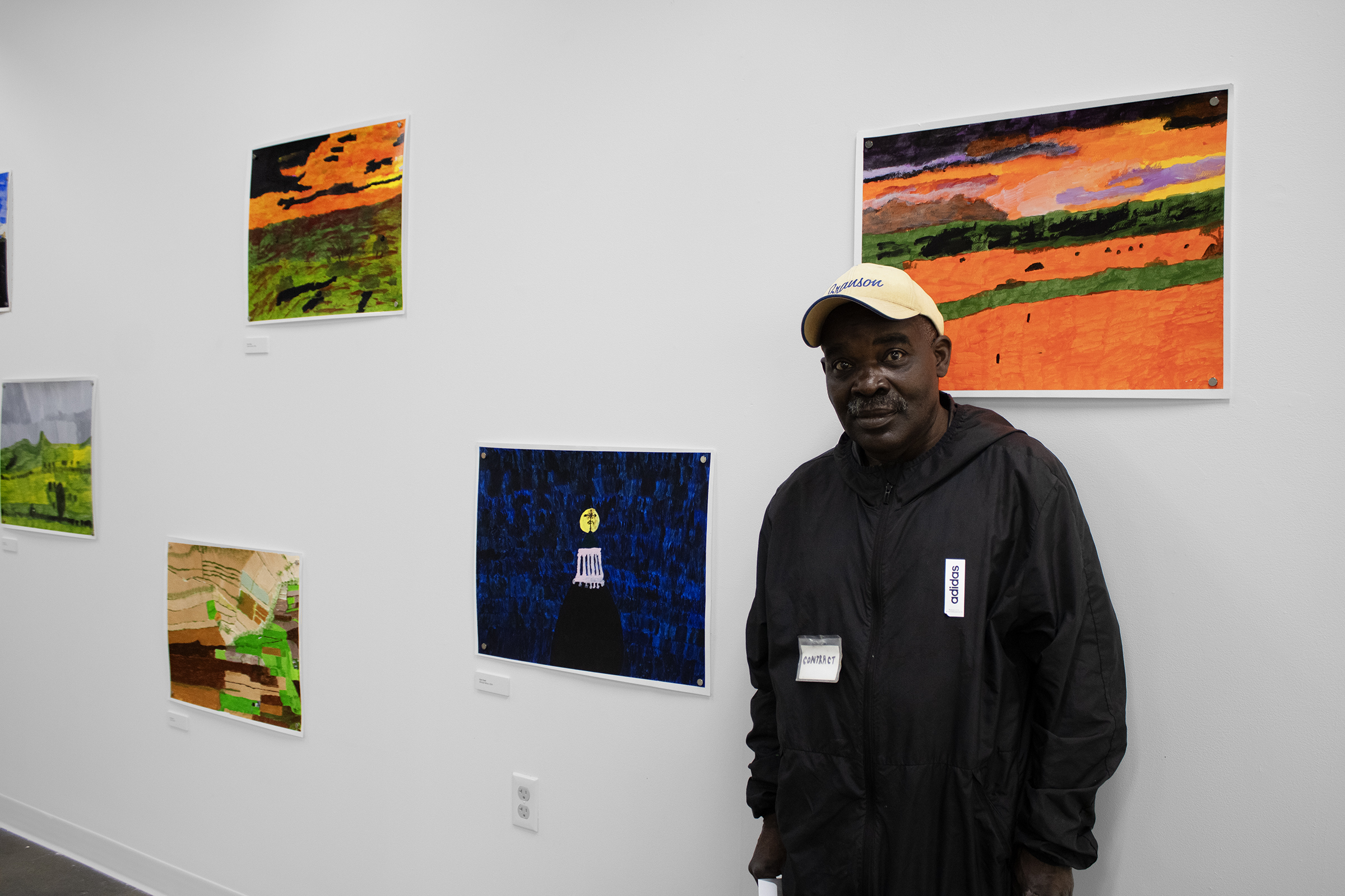 A man in a black jacket and yellow cap stands in an art gallery beside several framed landscape paintings.