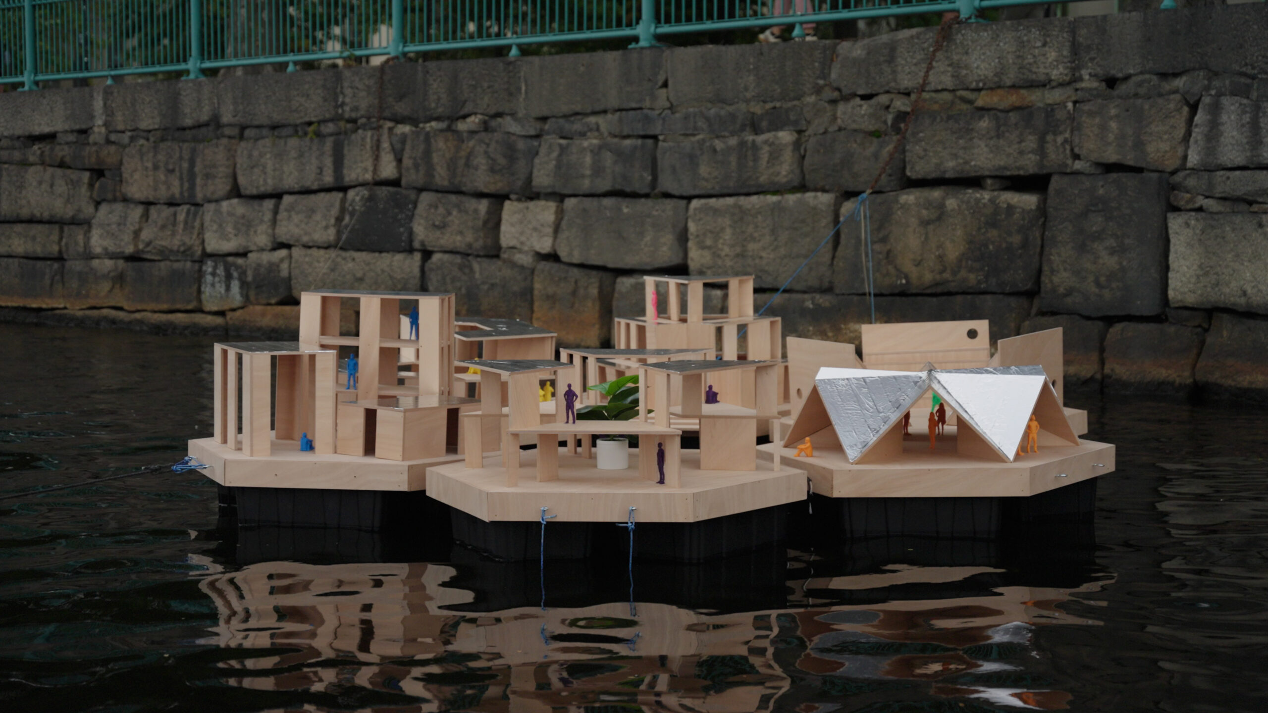 A miniature model of a floating wooden village from MIT Future Fest, featuring small plastic figures anchored in water against a stone wall.