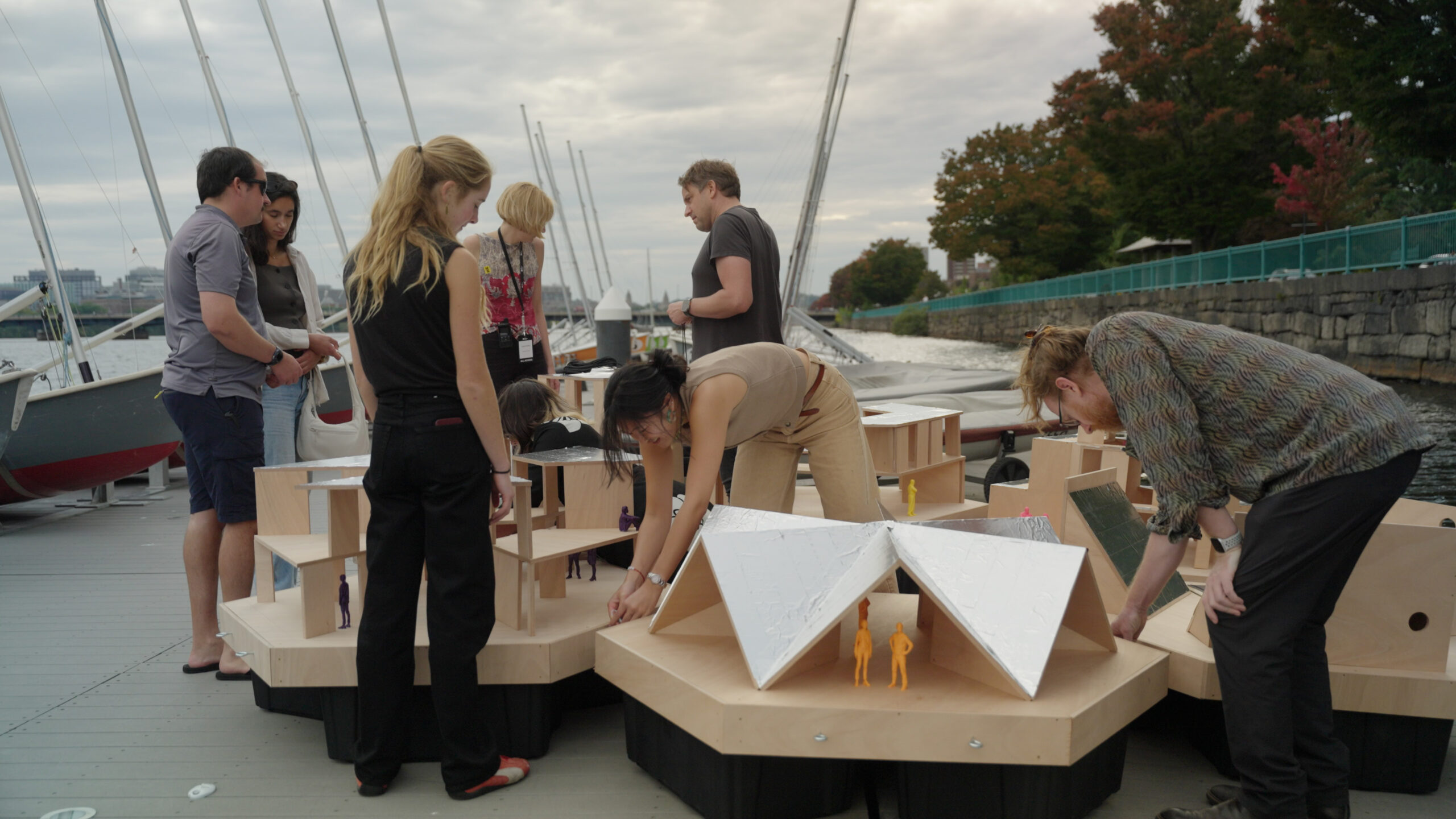 A group of people standing on a dock at MIT Future Fest, surrounding and interacting with several modular wooden architectural models.