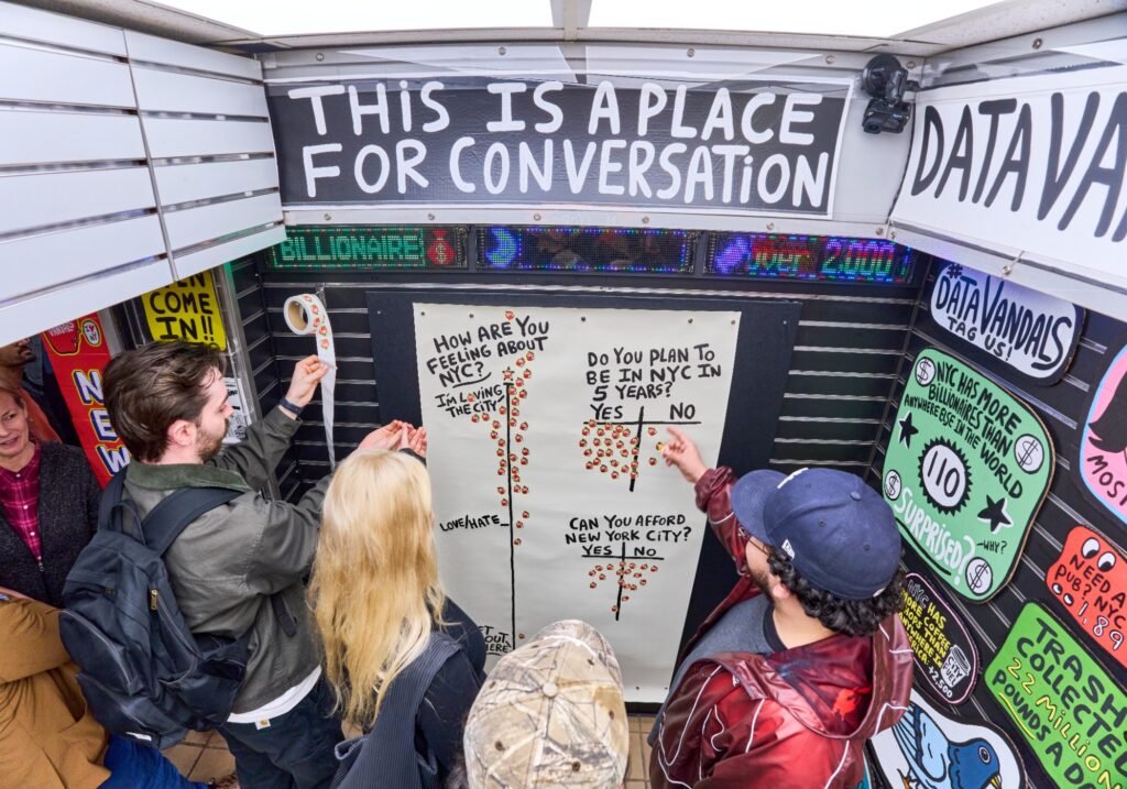 Four people place stickers on a board with survey questions inside a small booth labeled “This is a place for conversation” at Archtober 2026, surrounded by colorful posters and signs.