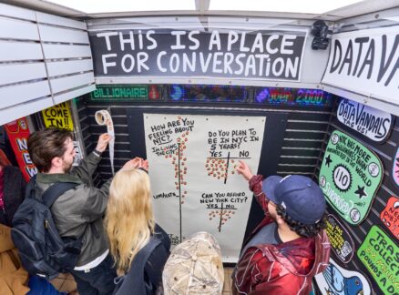 Four people place stickers on a board with survey questions inside a small booth labeled “This is a place for conversation” at Archtober 2026, surrounded by colorful posters and signs.