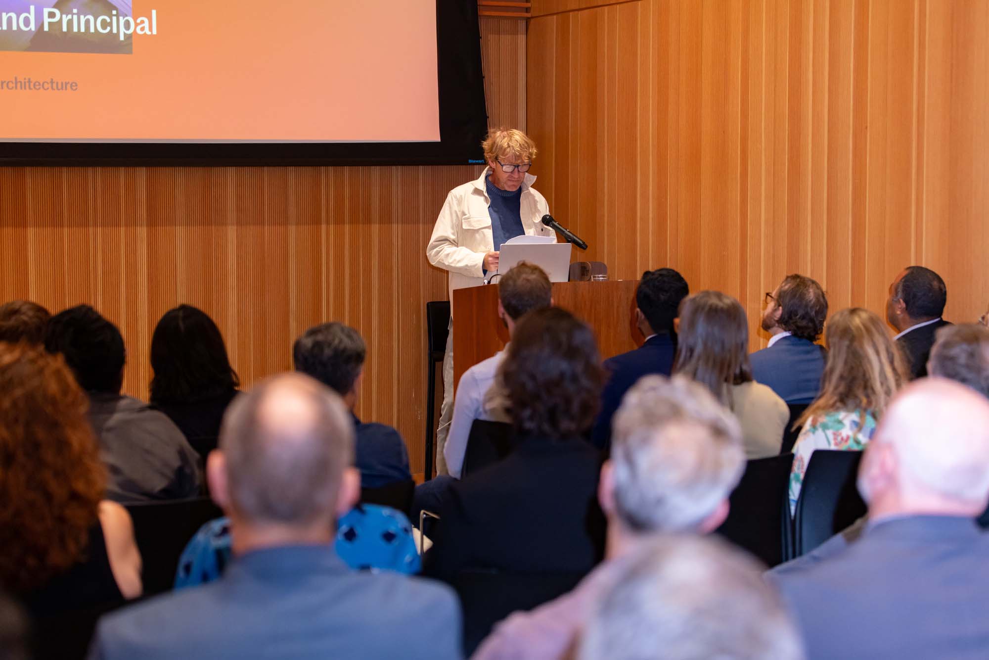A woman in a white jacket stands at a podium giving a presentation to an audience in a wood-paneled room.