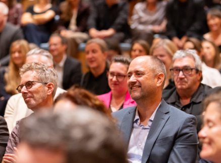 A diverse audience seated in an auditorium, with a smiling man in a suit jacket in the foreground.