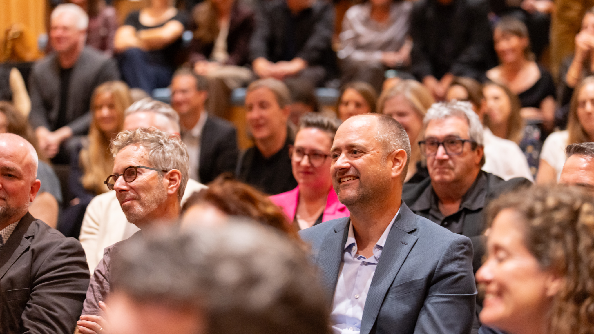 A diverse audience seated in an auditorium, with a smiling man in a suit jacket in the foreground.