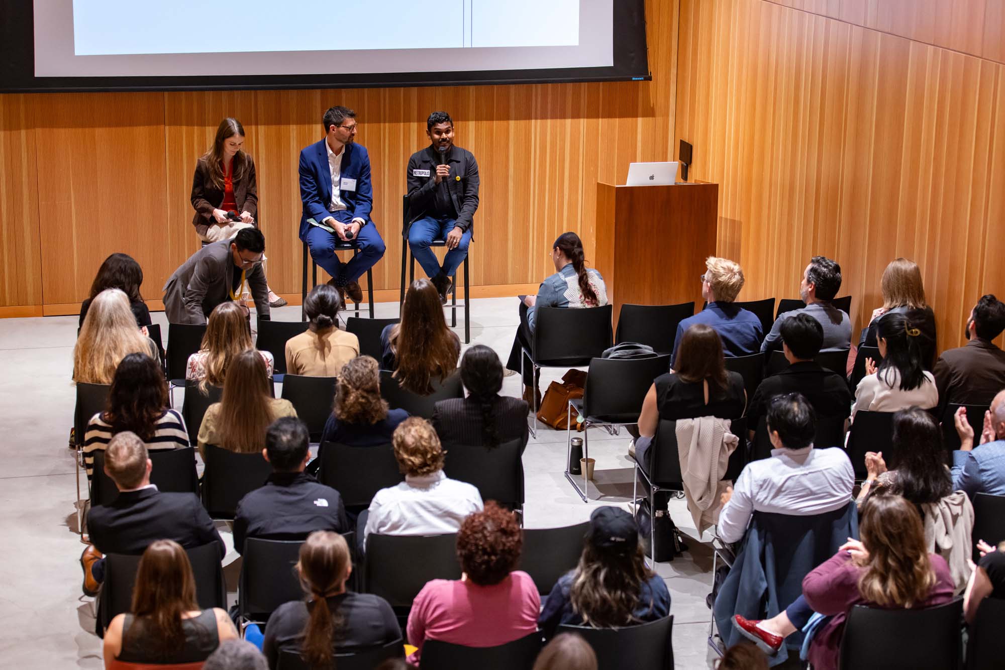 Three panelists sitting on stage at a conference, speaking to an audience seated in an auditorium.