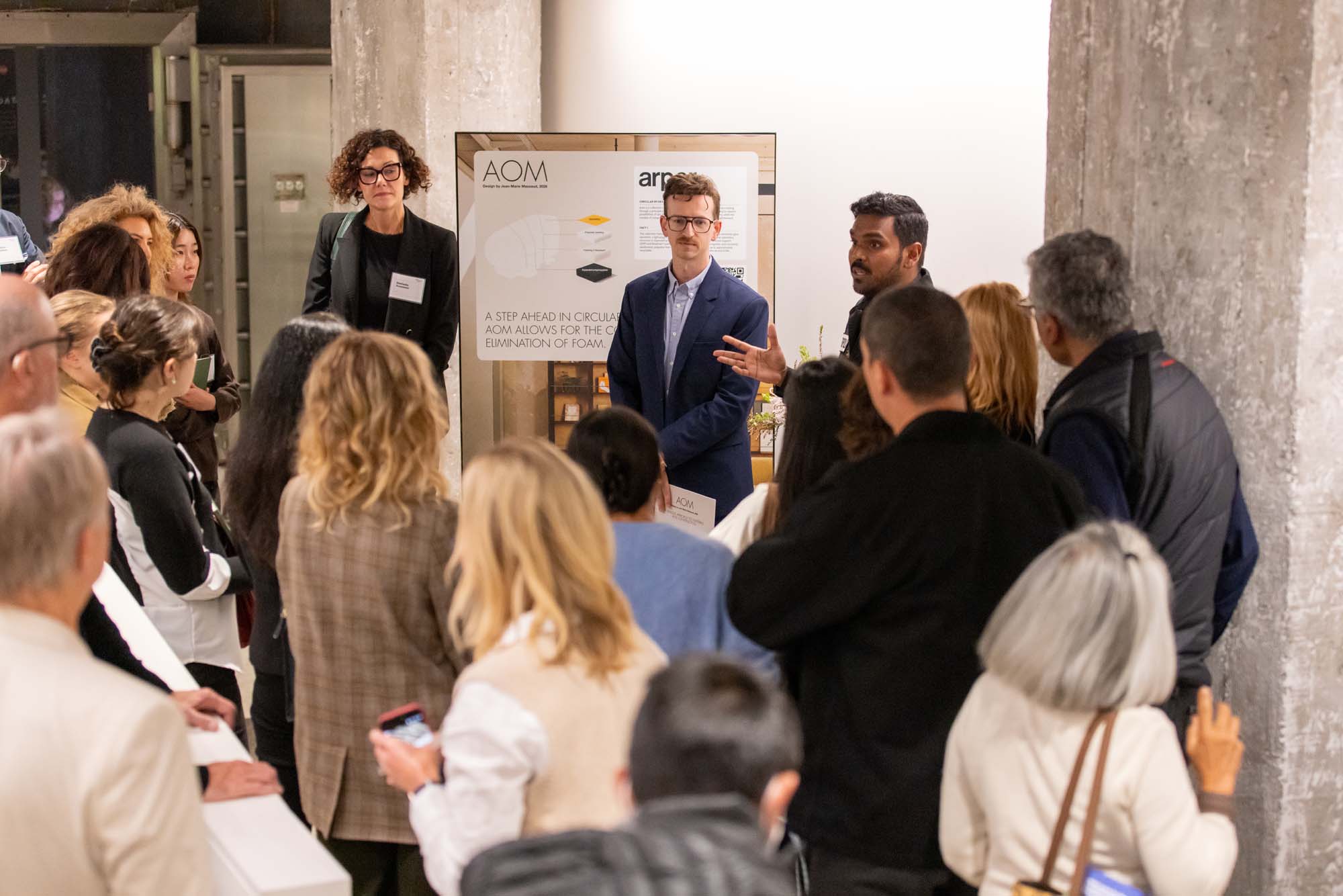 A man in a blue blazer stands before a group of people, gesturing toward an AOM project display board.