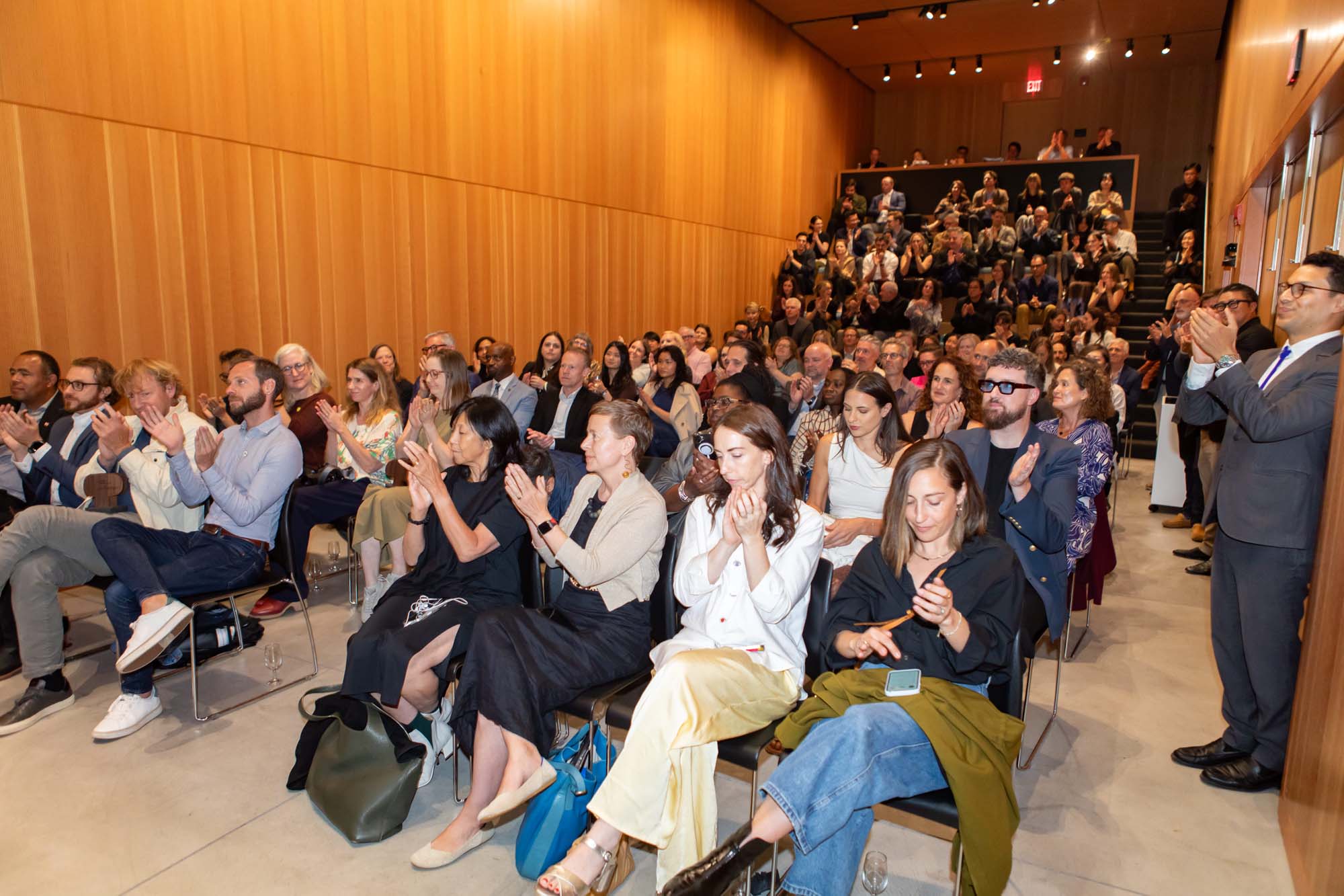 An audience seated in a wooden-paneled auditorium applauds during a presentation.