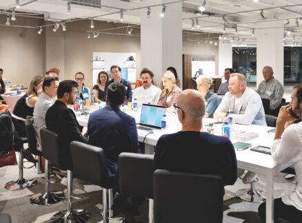 A group of people seated around a long white table in an office space, engaged in a discussion.