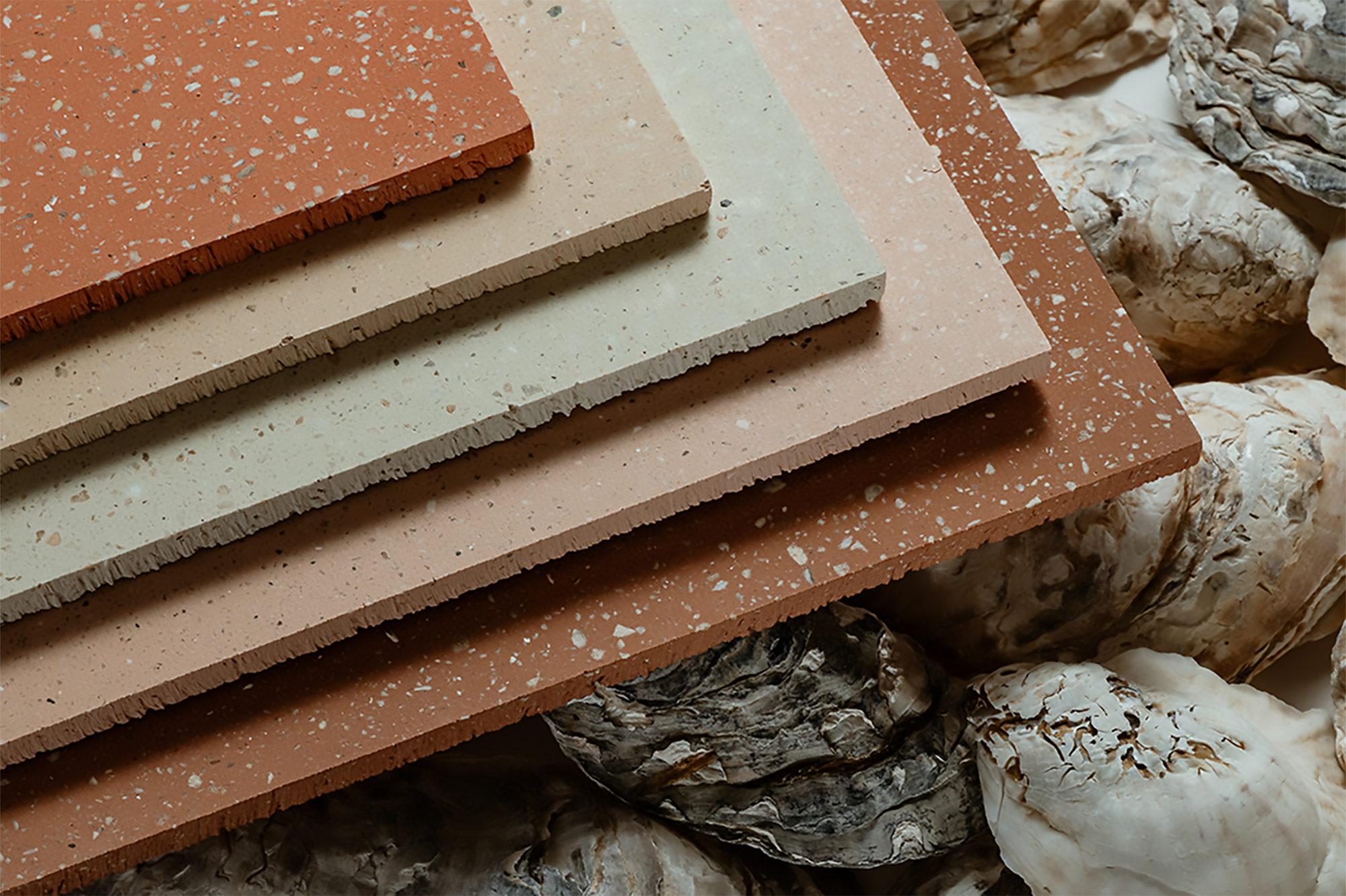 Four rectangular terrazzo tiles in earthy tones are stacked on top of oyster shells, showing their speckled surfaces and varying thicknesses.