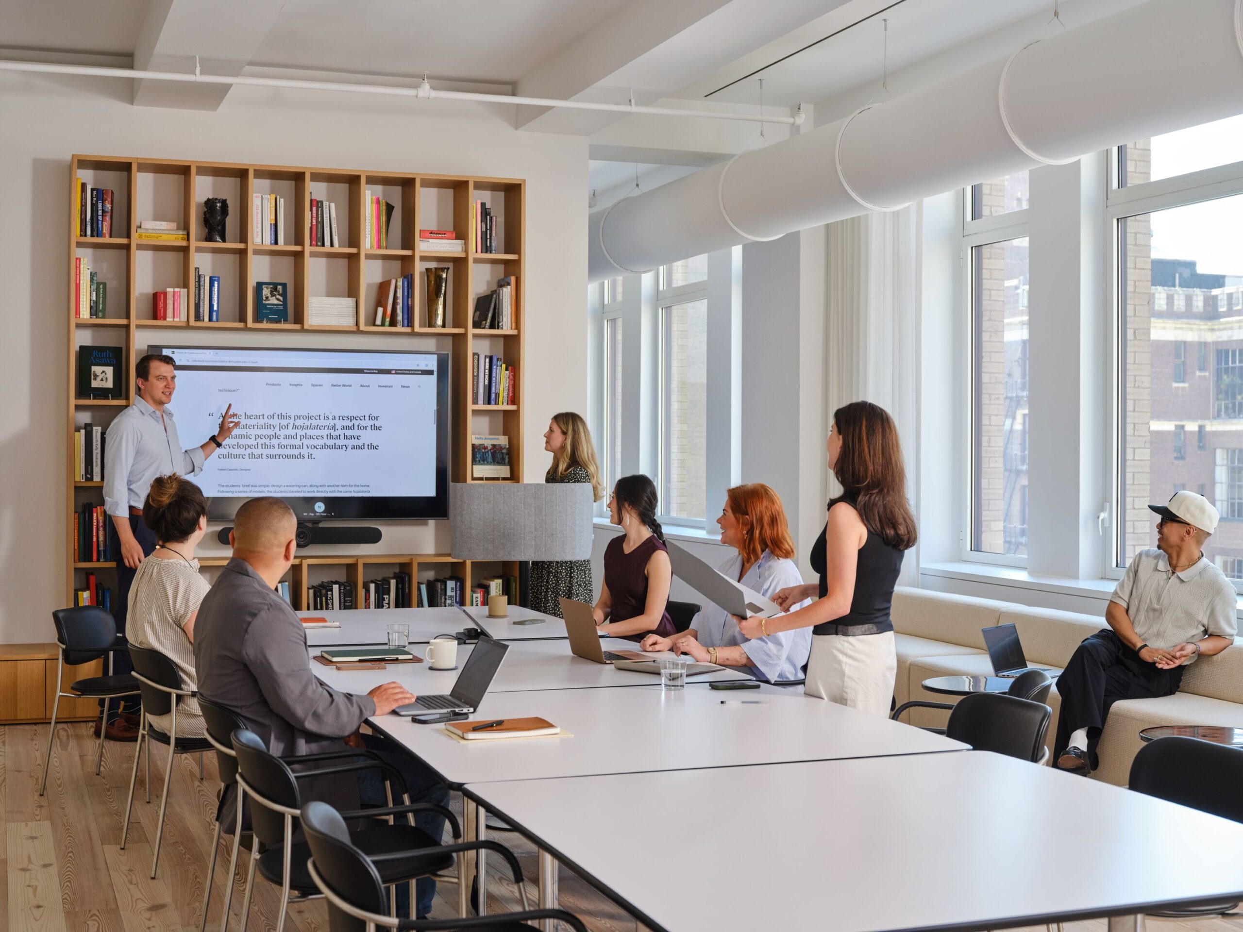 A man leads a presentation for a group of colleagues in a modern MillerKnoll conference room.