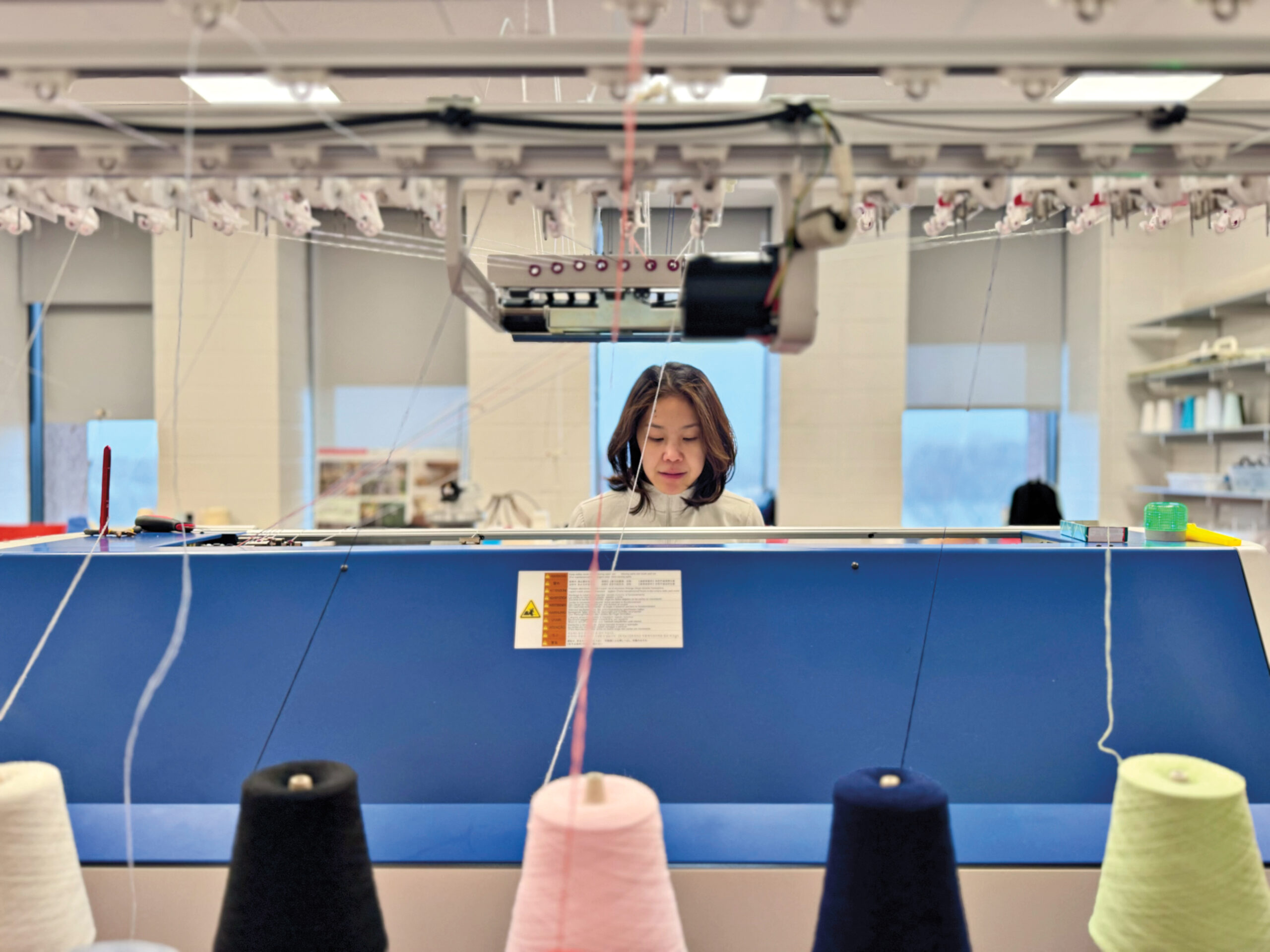 Spools of pink, green, navy, black and white thread feed into a 3D-printing machine, operated by a woman.