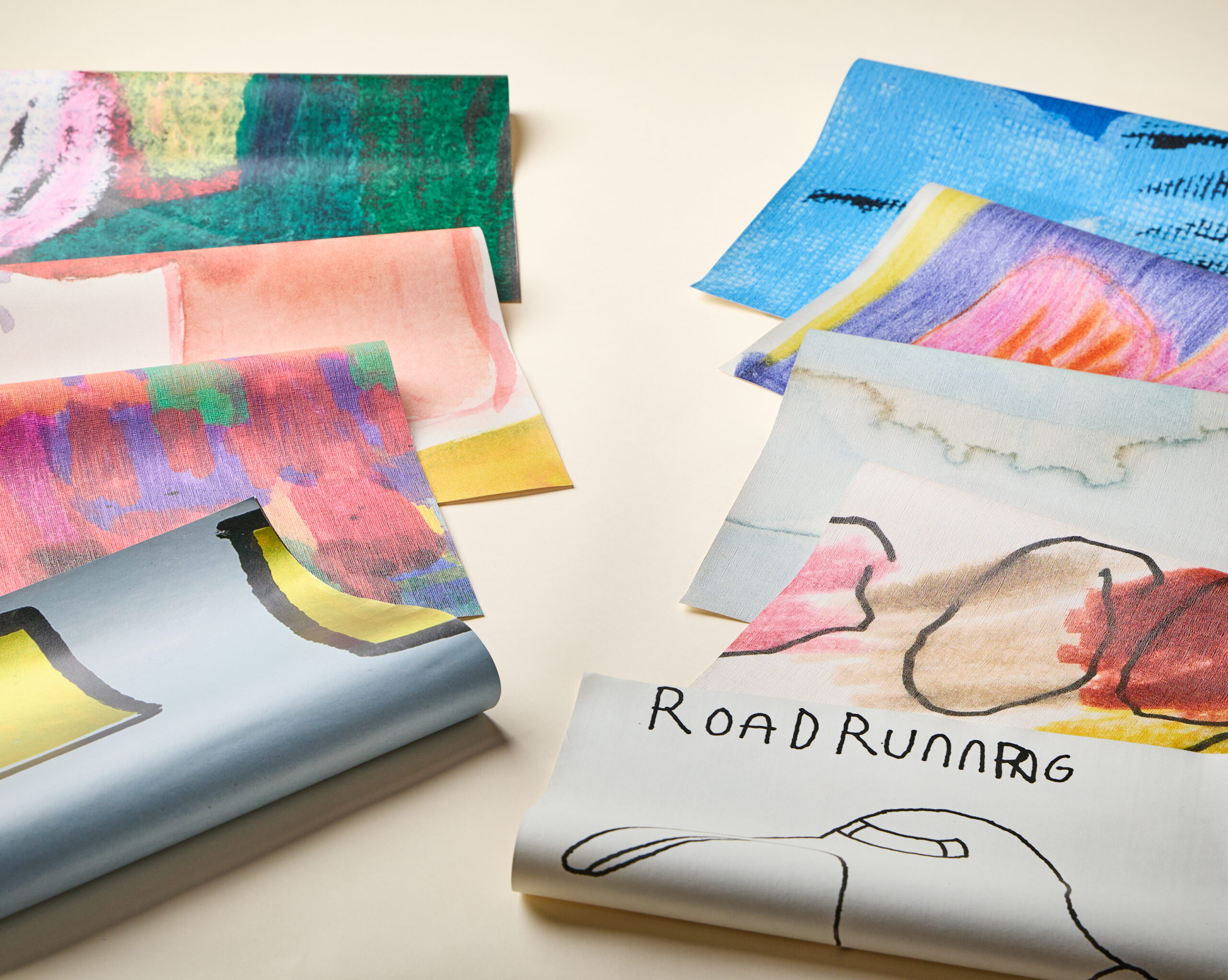 Several sheets of abstract, colorful art paper arranged on a light surface, with one sheet featuring the hand-drawn text “ROADRUNNER” and a line-art bird.