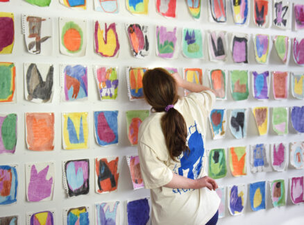 A person with long brown hair stands facing a wall covered in rows of colorful abstract watercolor paintings.