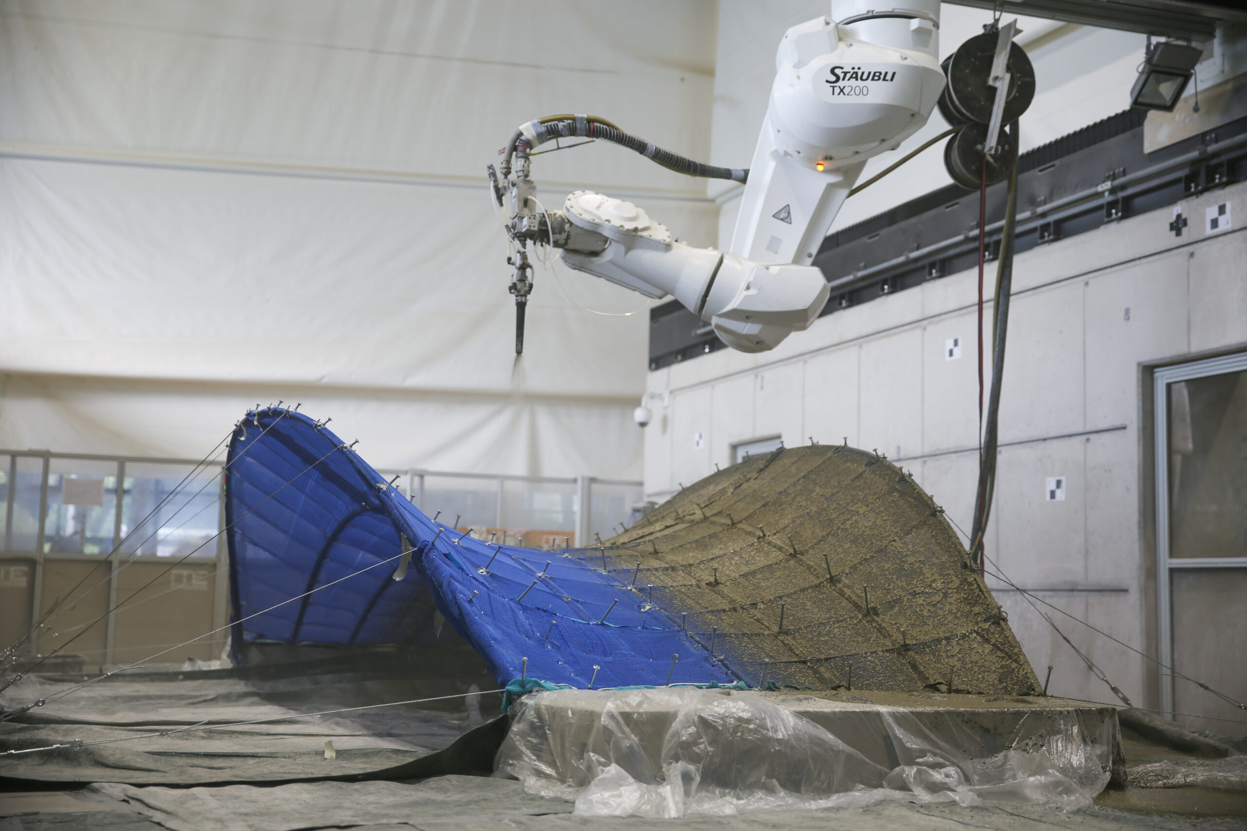 A robotic arm sprays material onto a large, curved structure partially covered in blue mesh during MIT Future Fest.