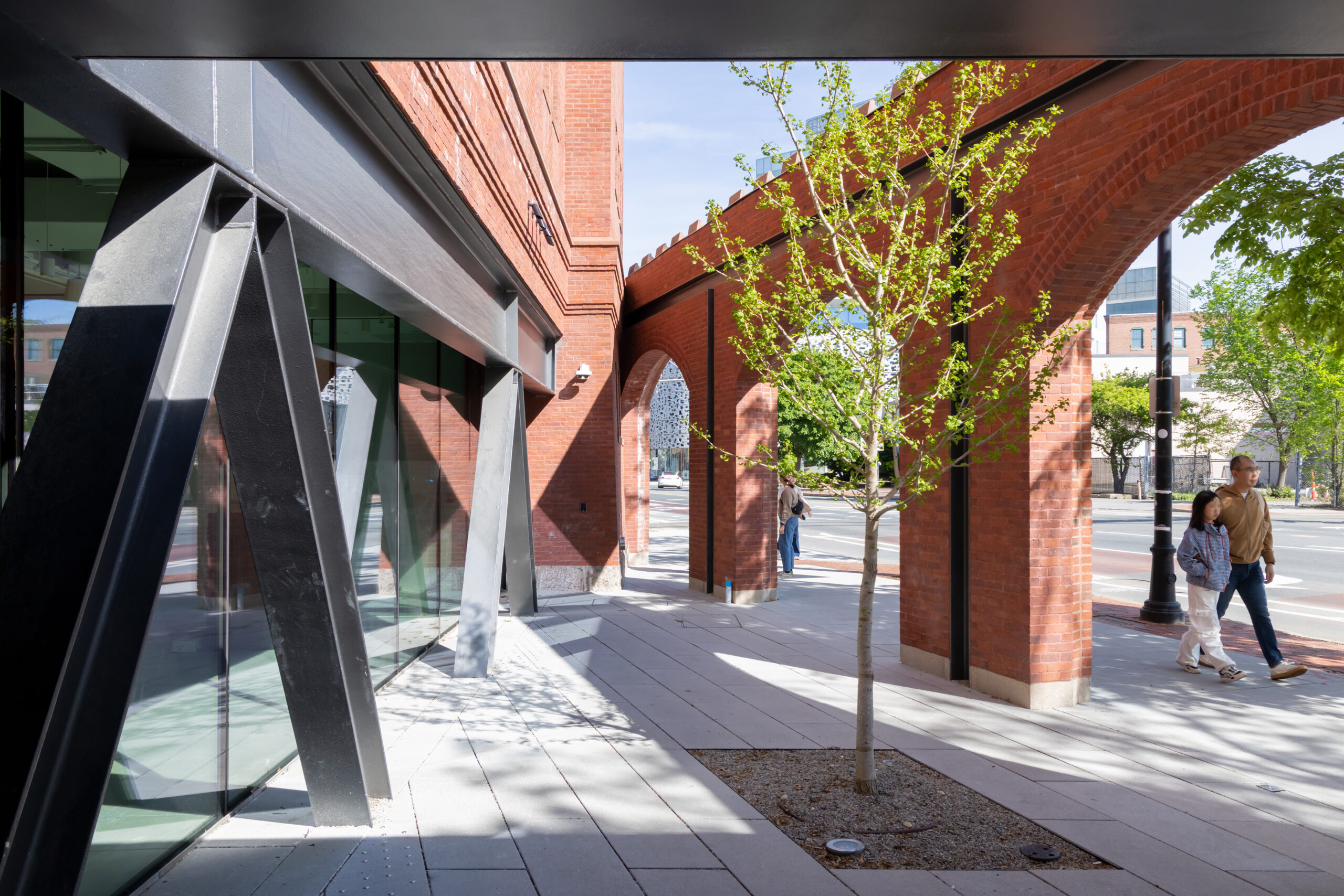 A brick building with arched walkways, a glass storefront with black steel supports, and two people walking on a paved plaza at MIT Future Fest.