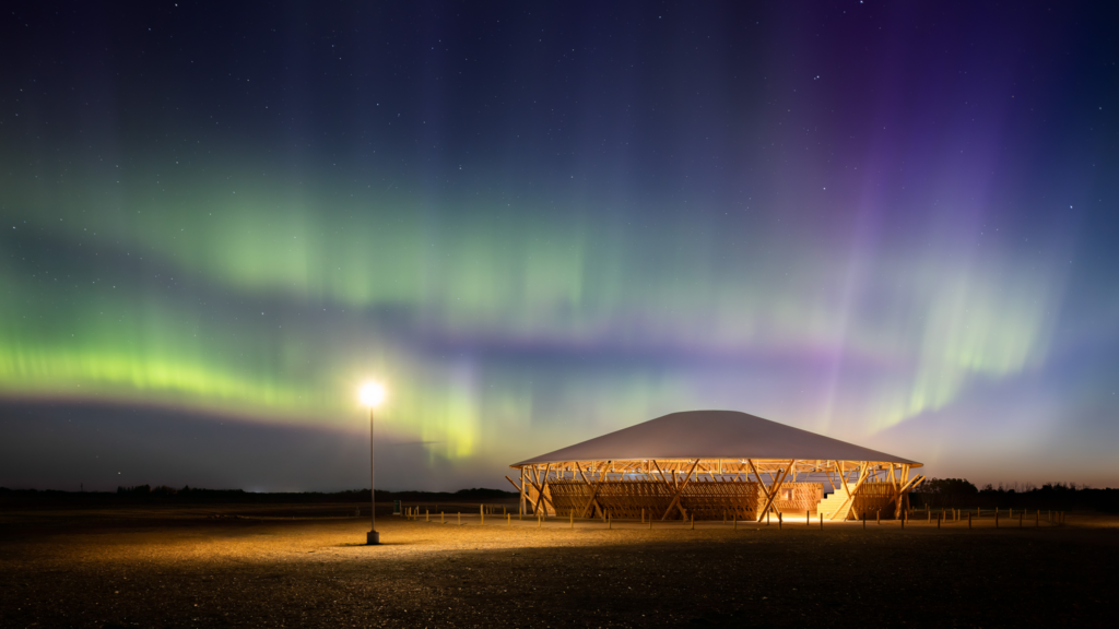 Aurora borealis in the night sky above a lit, circular wooden pavilion.