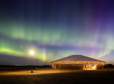 Aurora borealis in the night sky above a lit, circular wooden pavilion.