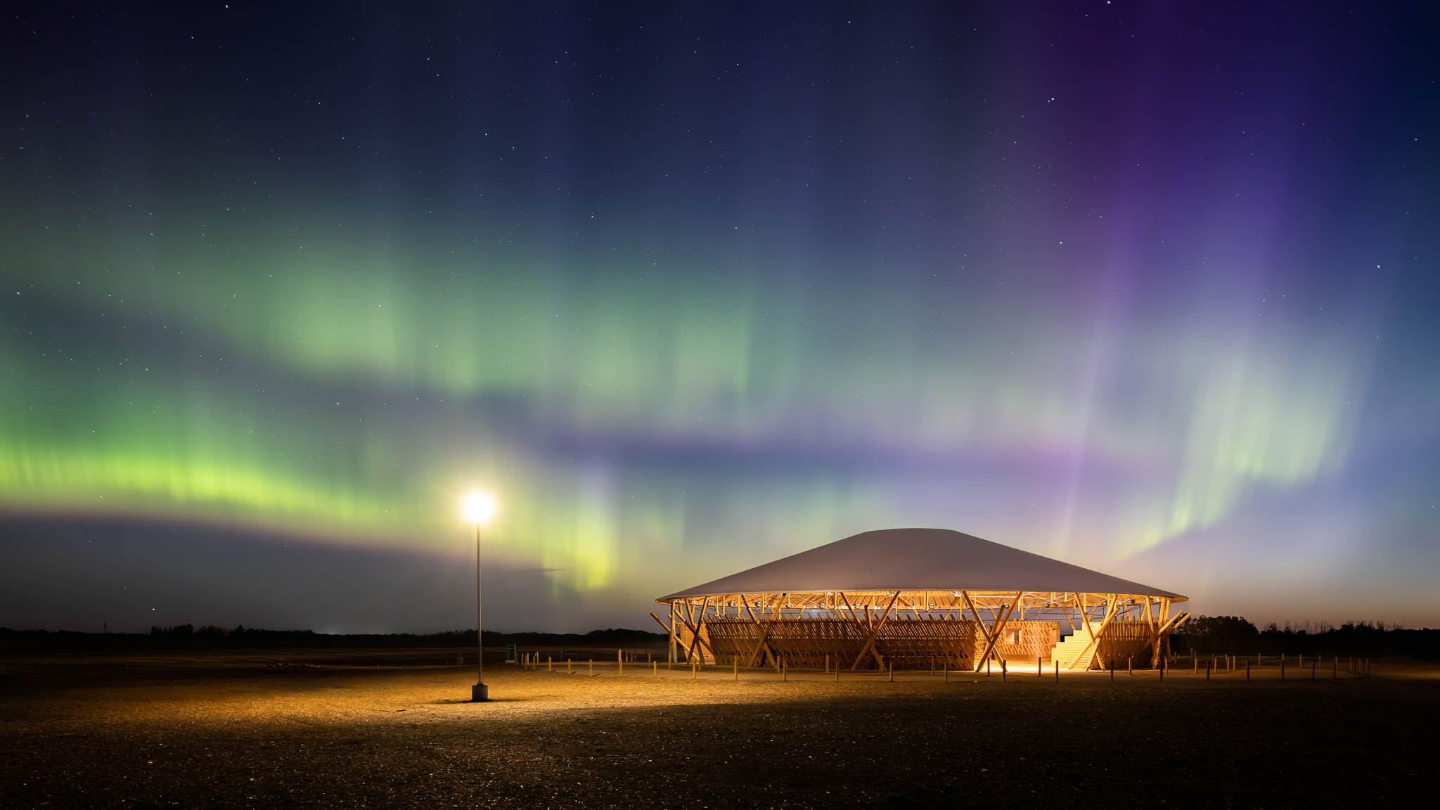 Aurora borealis in the night sky above a lit, circular wooden pavilion.