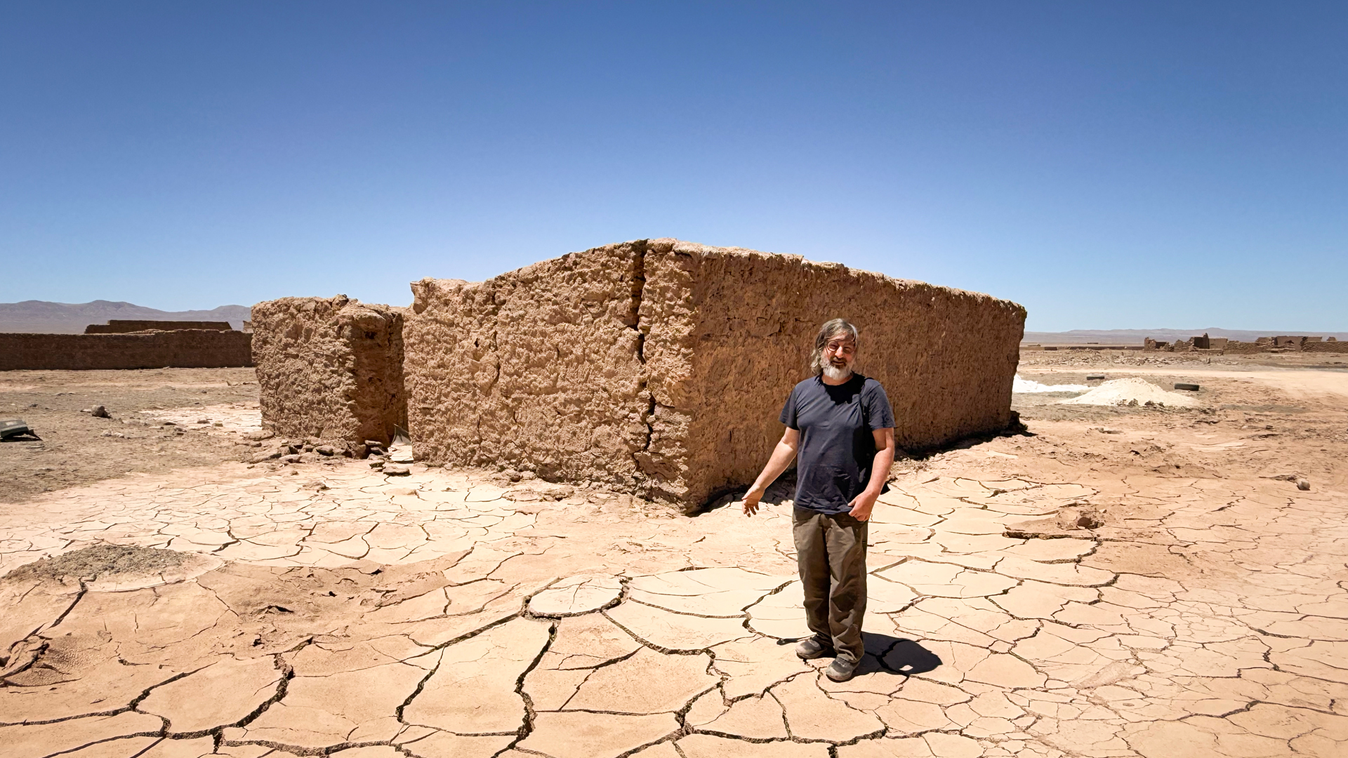 A man stands on cracked, dry earth next to the ruins of an adobe building in an arid Archtober 2026 landscape.