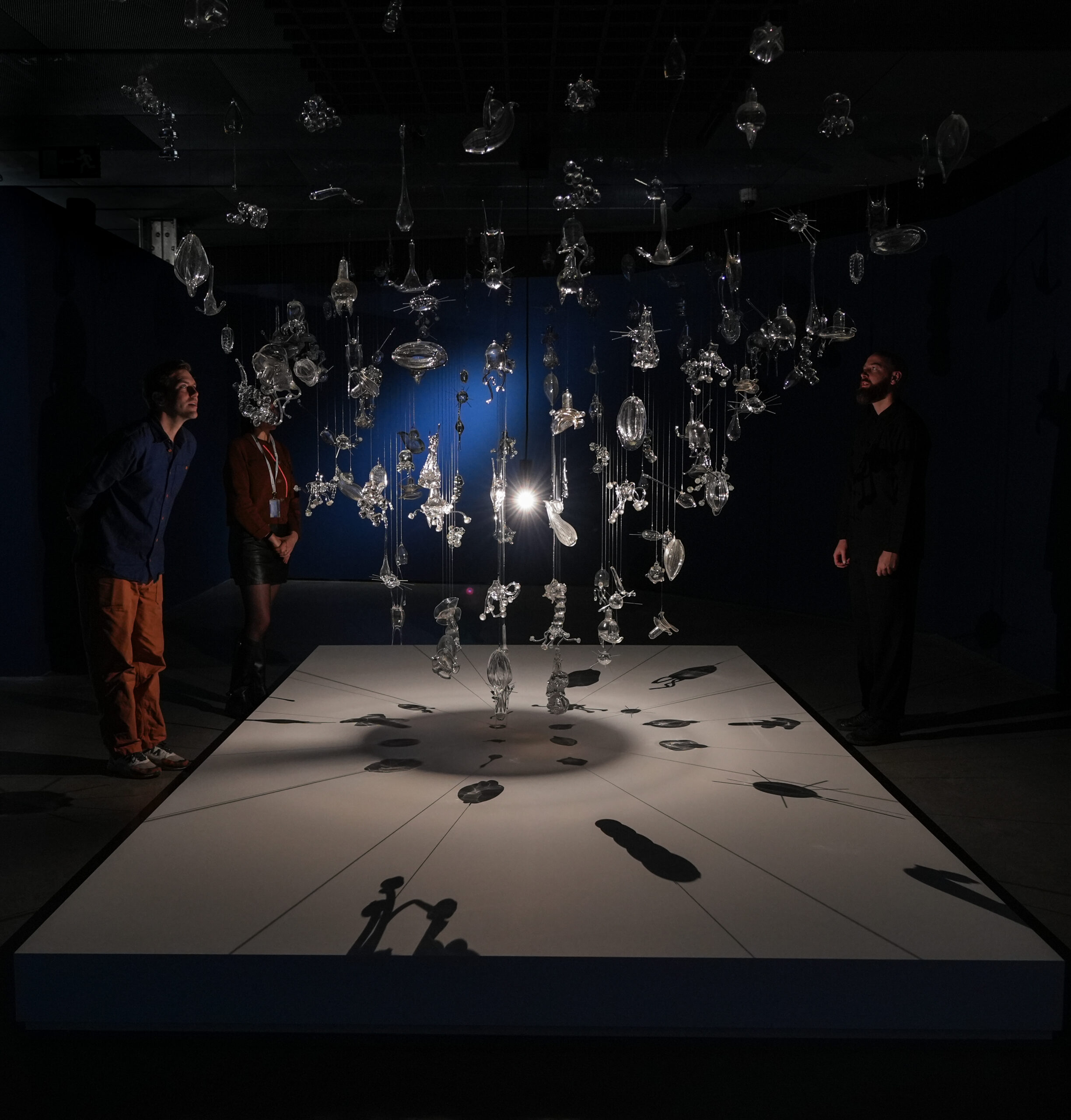 Three people stand at MIT Future Fest around a platform displaying a hanging cluster of clear glass sculptures that cast detailed shadows onto the surface below.