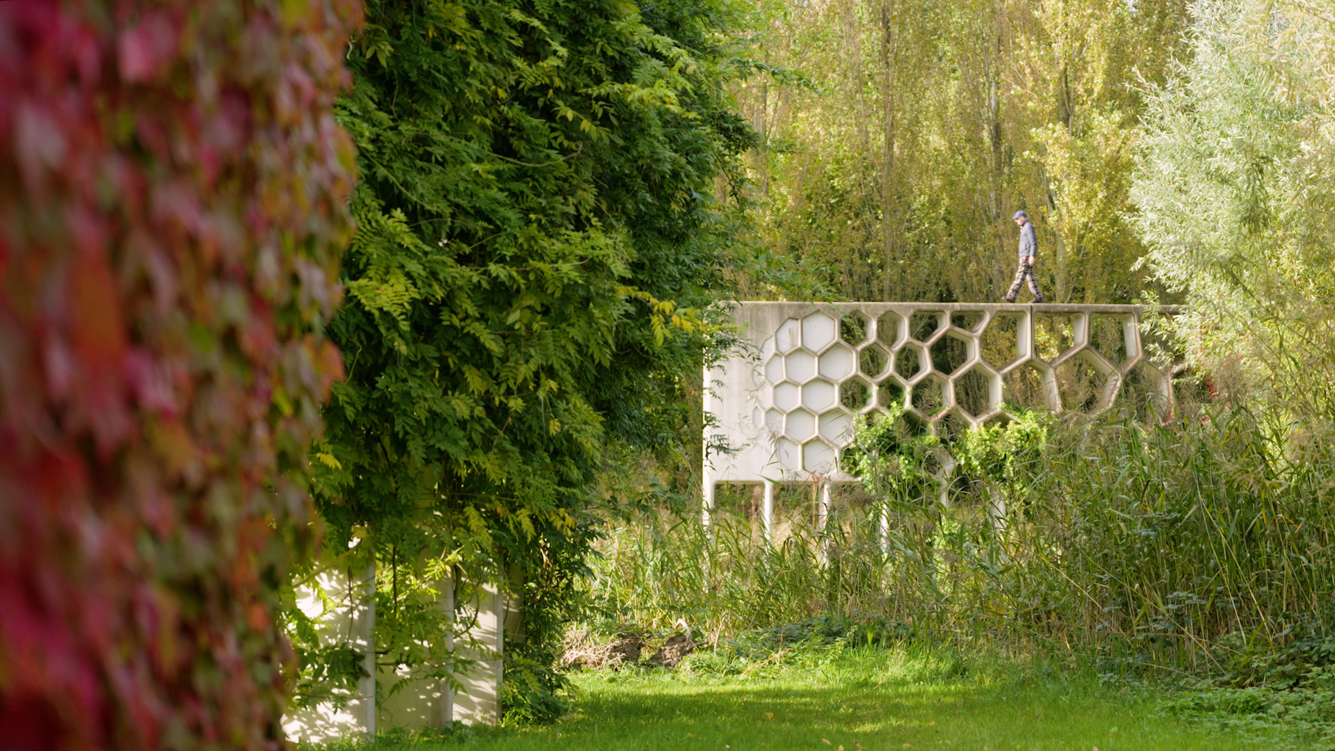 A person walks along the top of a concrete wall with a hexagonal cutout pattern in an Archtober 2026 park surrounded by trees and foliage.