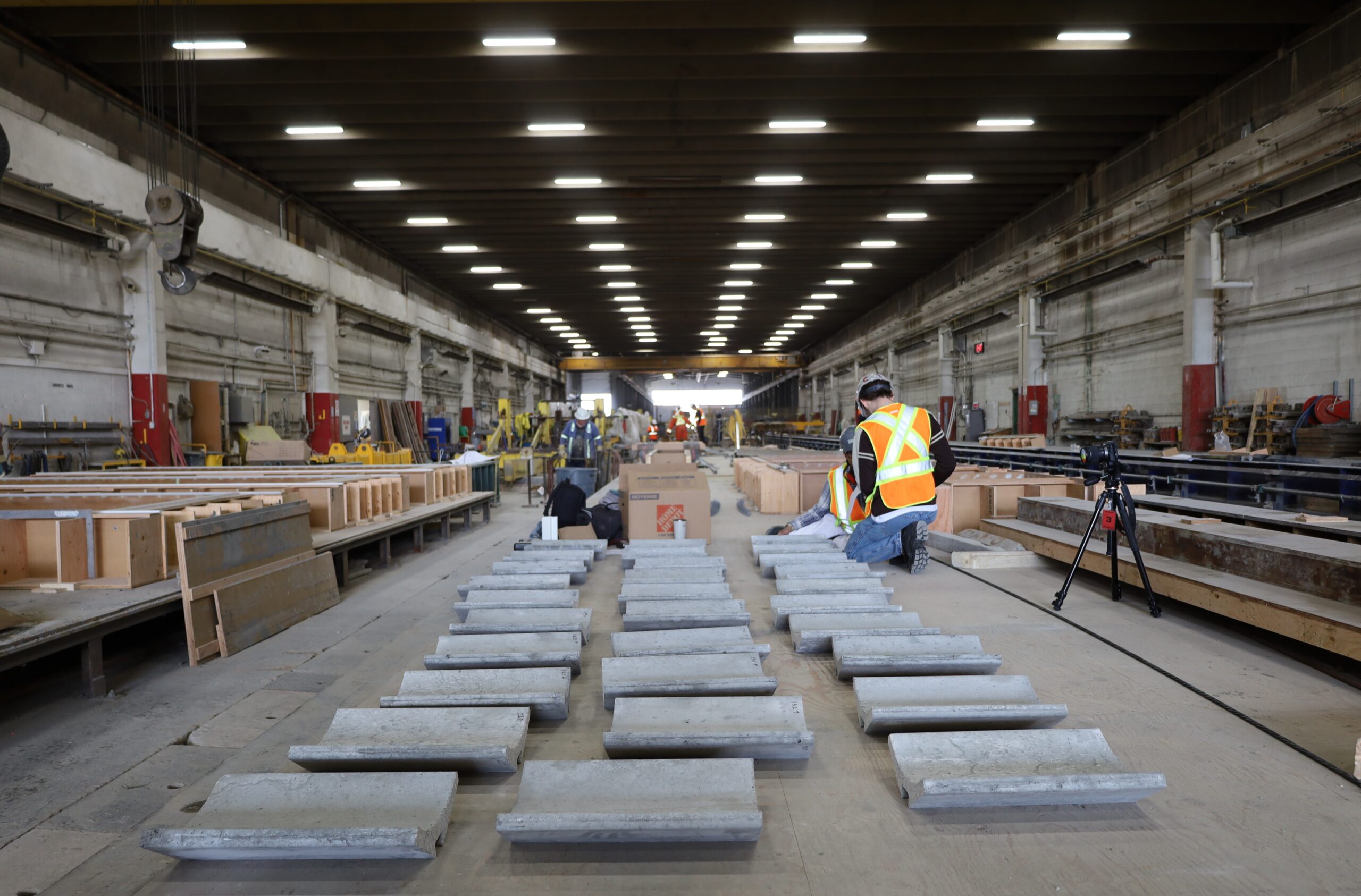 Workers in high-visibility vests inspect rows of concrete drainage channels in a large warehouse at MIT Future Fest.