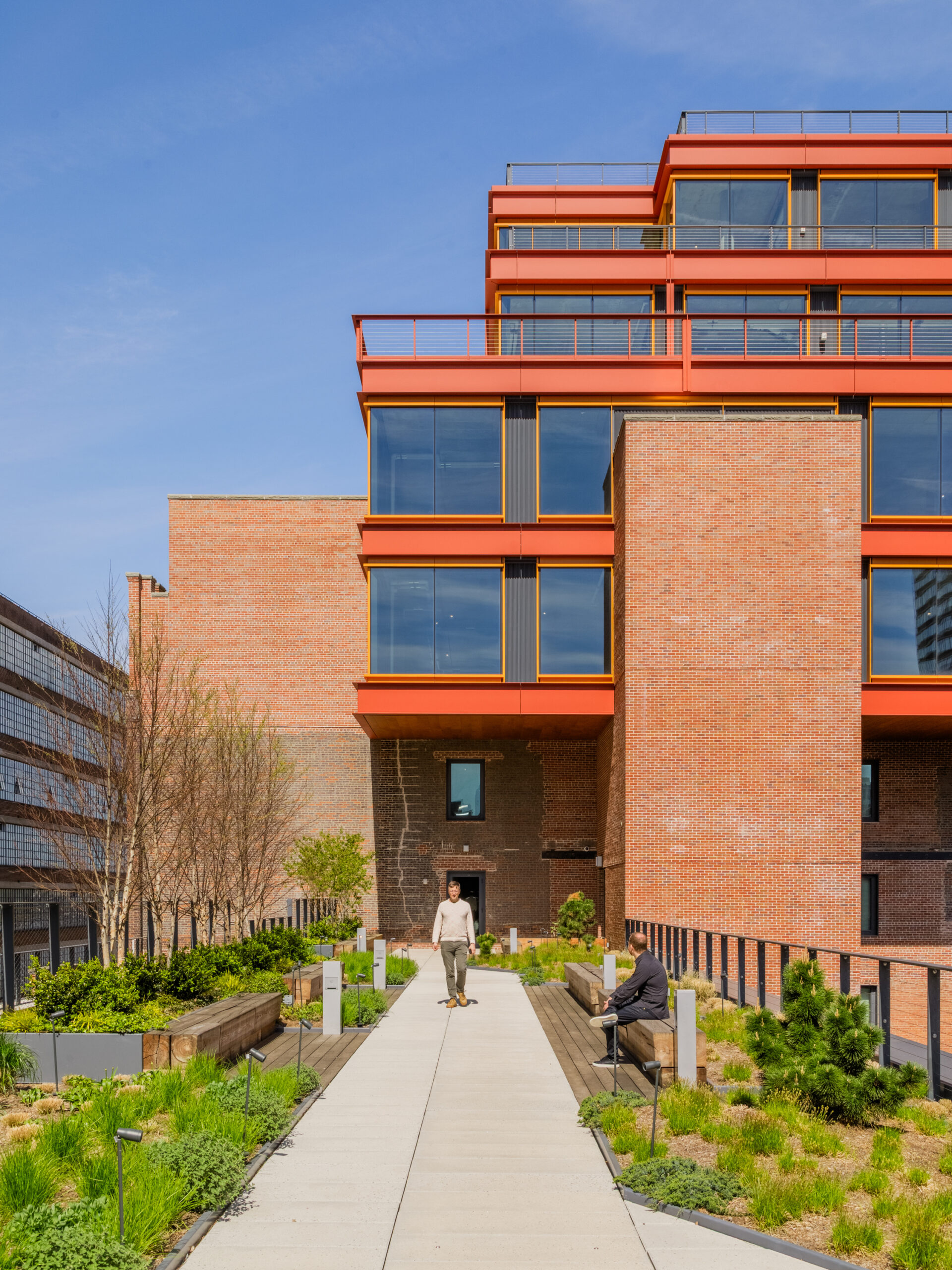 A paved walkway in a biomimetic rooftop garden connects a modern orange-framed building to an older brick structure, with one man walking along the path and another sitting on a wooden bench.