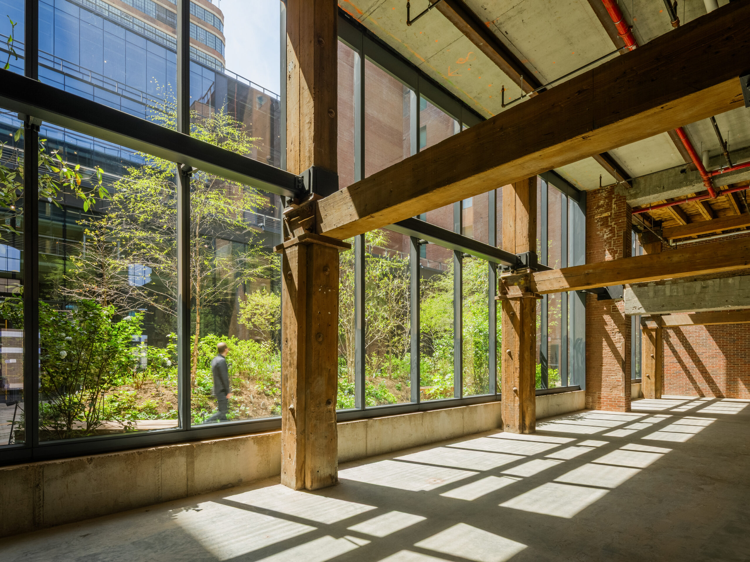 An empty industrial space with large windows overlooking a courtyard, featuring exposed timber beams, brick walls, and a person walking outside in a setting that evokes biomimicry.