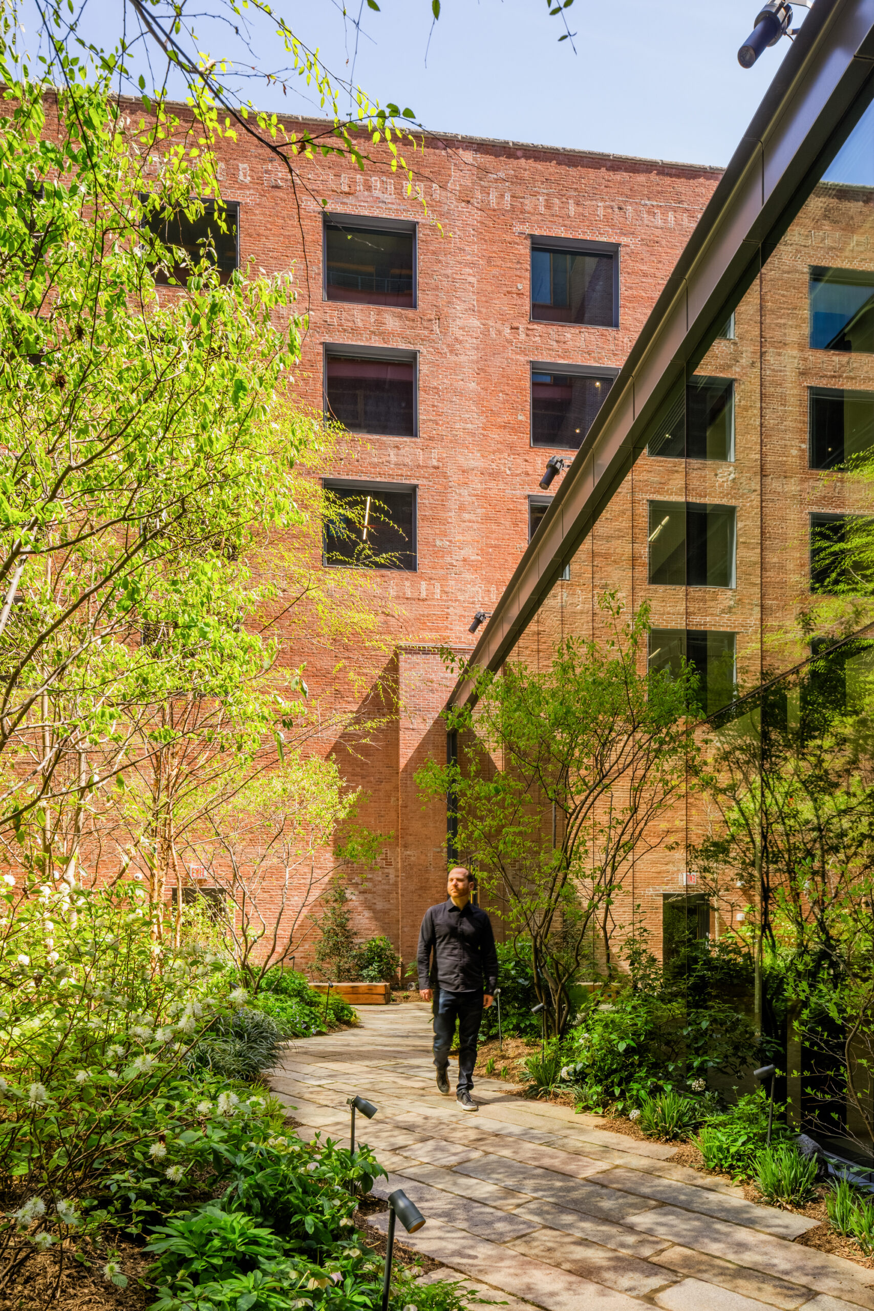 A person walks along a stone path in a courtyard between a brick building and a glass-walled structure reflecting biomimicry, surrounded by trees and shrubs.
