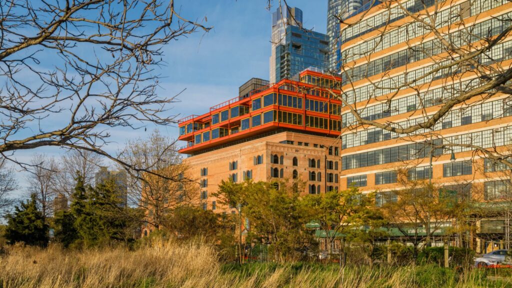 A multi-story brick building with a modern, biomimicry-inspired orange rooftop addition, viewed behind trees and tall grass.